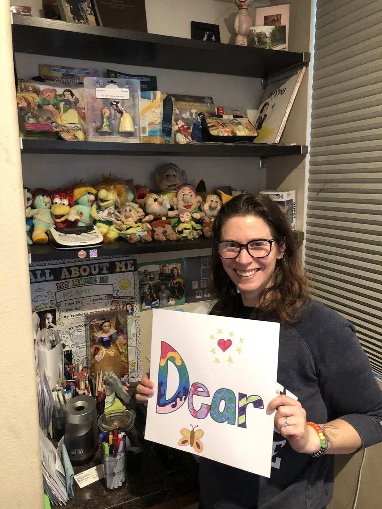 A woman with glasses and brown hair smiling and holding a colorful sign that says 'Dear' with a rainbow, heart, and butterfly illustrations. She is in a room with shelves filled with toys, plush puppets, and a desk with stationery and photos.