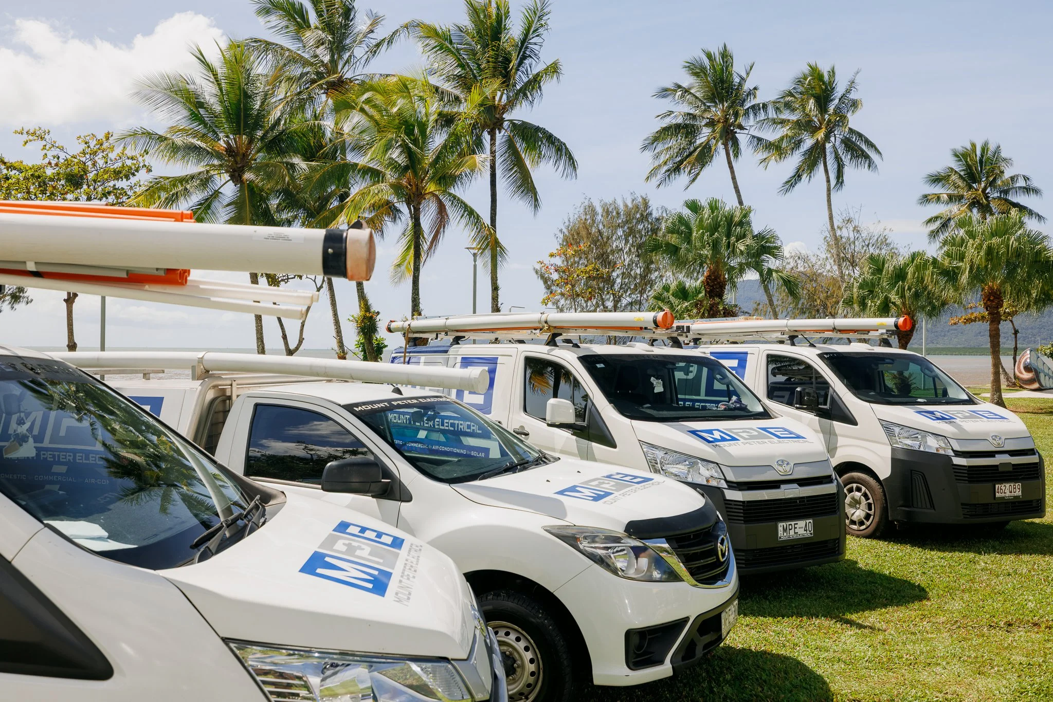White service trucks with ladders parked on grass near palm trees and a body of water in the background.
