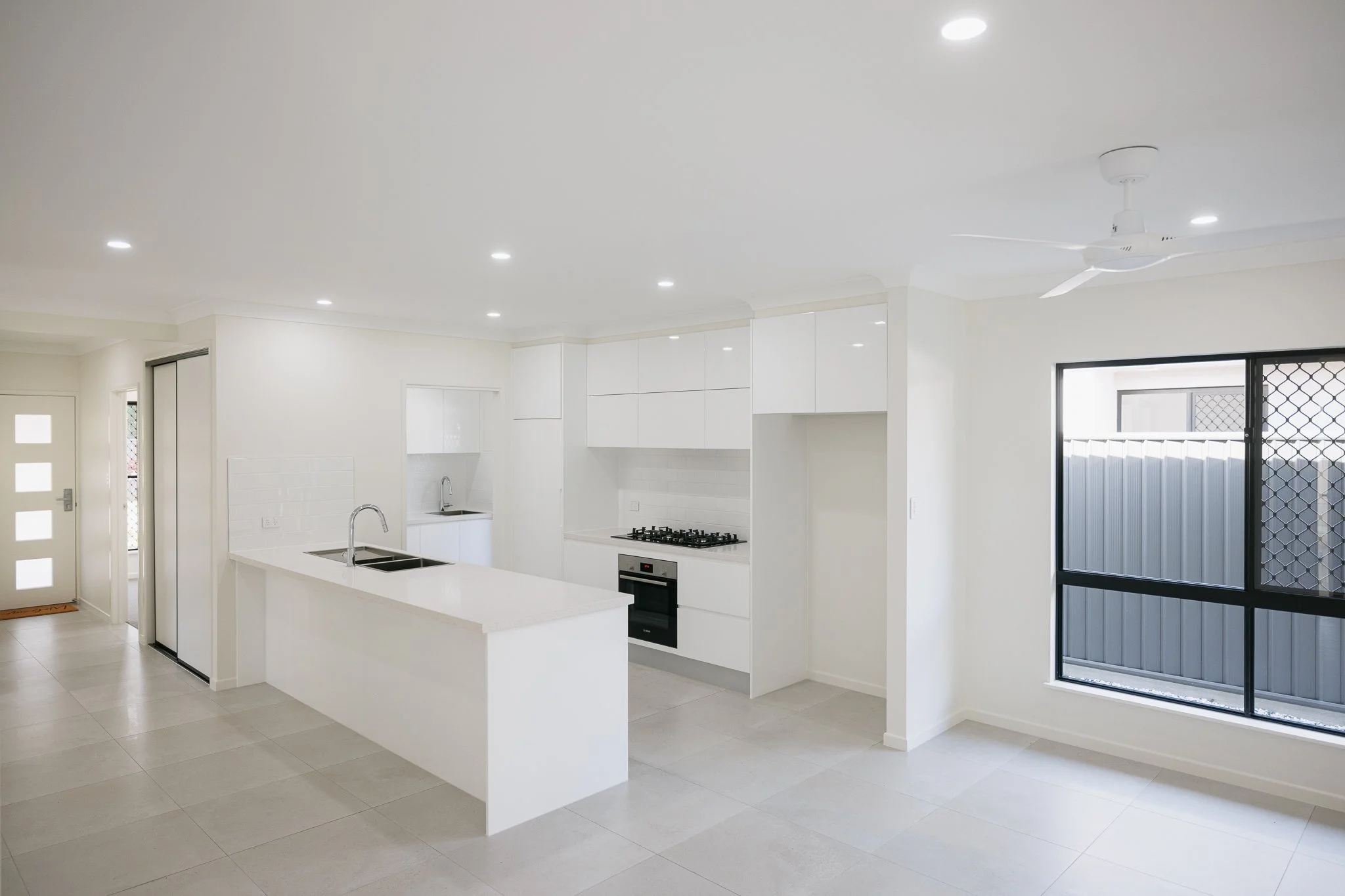 A modern, minimalist kitchen with white cabinets, a white island with a sink, a built-in oven, a gas stove, tiled backsplash, a ceiling fan, recessed lighting, and a sliding glass door with security screen.