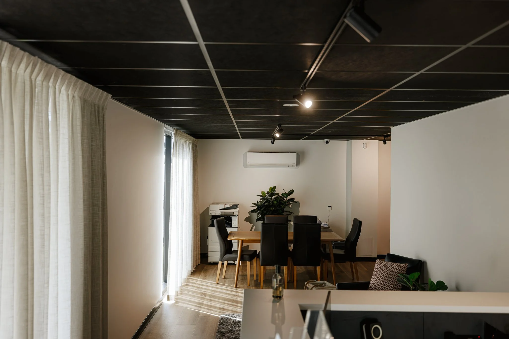 Modern living room with black ceiling panels, white walls, a dining area with a wooden table and black chairs, large windows with curtains, a potted plant, and an air conditioning unit.
