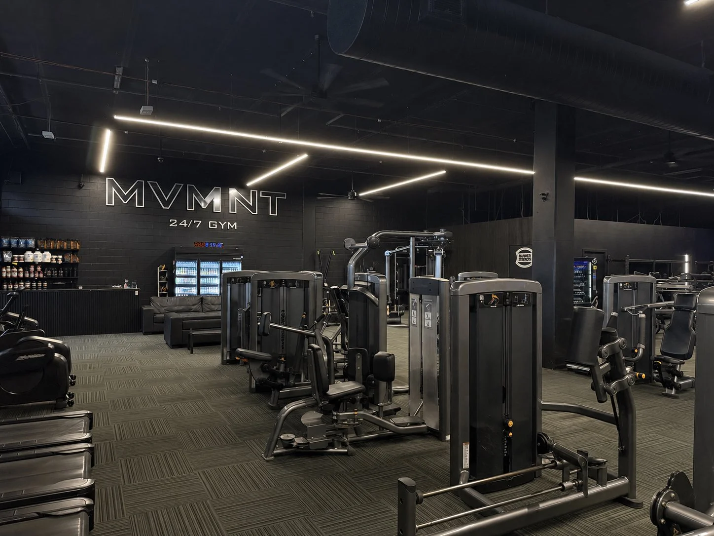 Interior of a modern, 24/7 gym with fitness equipment, black walls, and LED lighting, with a sign reading 'MVMT 24/7 GYM' on the wall.