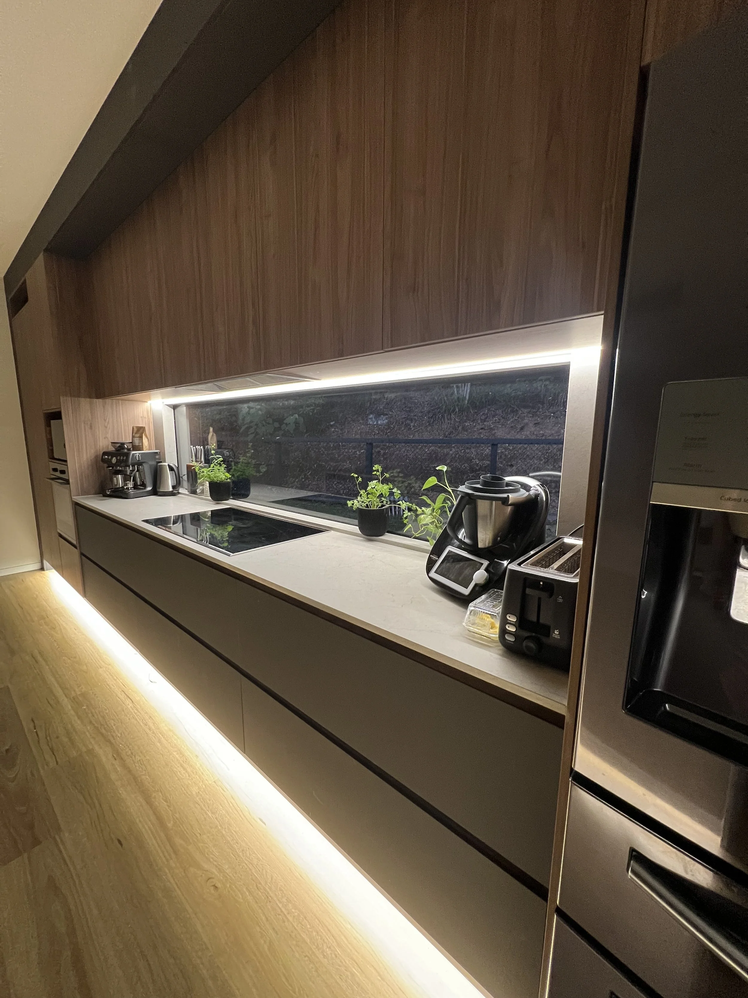 Modern kitchen with wooden cabinets, white countertop, large window, plants, coffee machine, toaster, and kitchen appliances, illuminated by under-cabinet lighting and warm floor lighting.