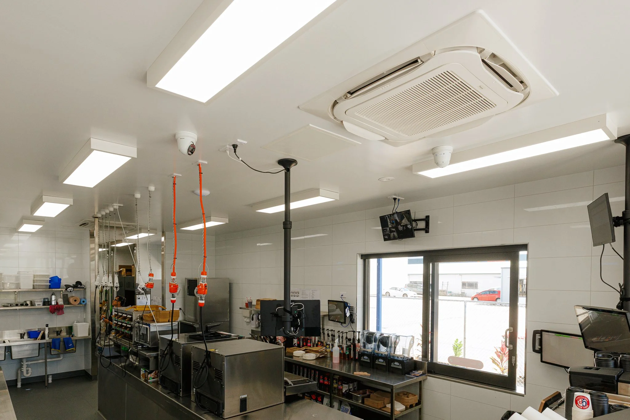 Interior of a clean commercial kitchen with ceiling lights, a ceiling-mounted air conditioning unit, security cameras, and various kitchen equipment and supplies on shelves and counters.