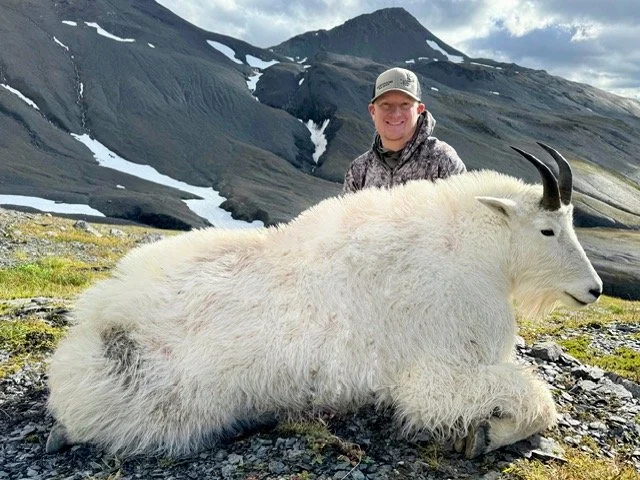 A smiling man with a hat kneeling next to a large, Kodiak mountain goat with black horns, on rocky terrain with snow-capped mountains in the background. Guided by Jason Bunch of Kodiak Backcountry Adventures