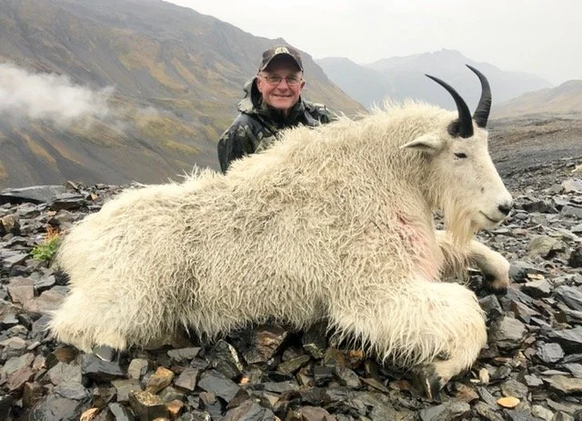 A man in outdoor gear smiling behind a large, white goat with black horns lying on rocky terrain with a mountain landscape in the background. Guided by Jason Bunch of Kodiak Backcountry Adventures