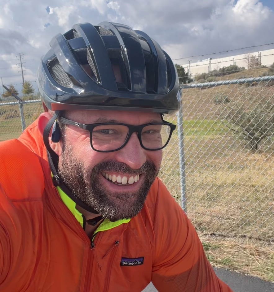 A man with glasses and a beard smiling while wearing a black bicycle helmet and an orange jacket, outdoors with a chain-link fence and cloudy sky in the background.