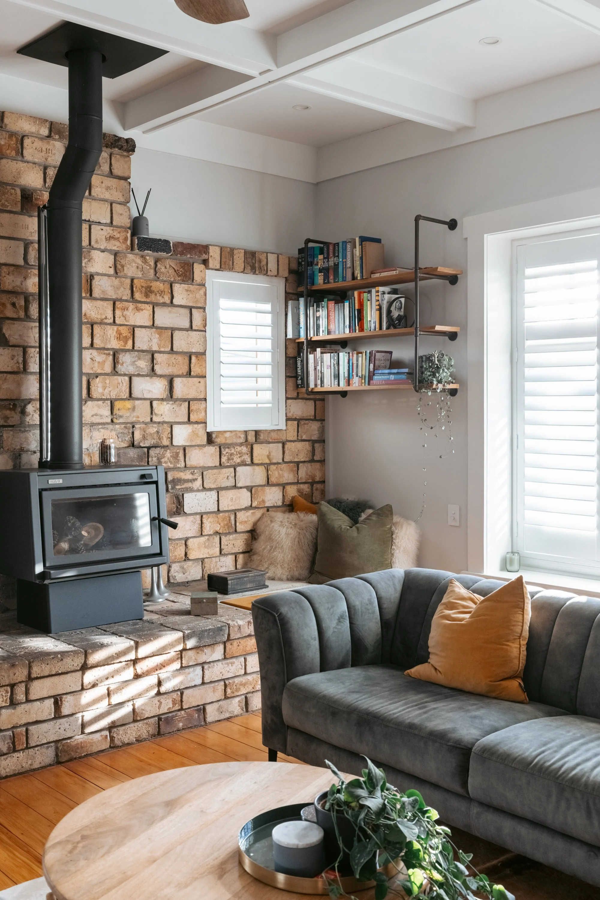Living room with brick fireplace, wood stove, bookshelf, gray sofa, orange pillow, and window with blinds. 1930's Bungalow renovation in Papamoa, New Zealand by BCN Studio