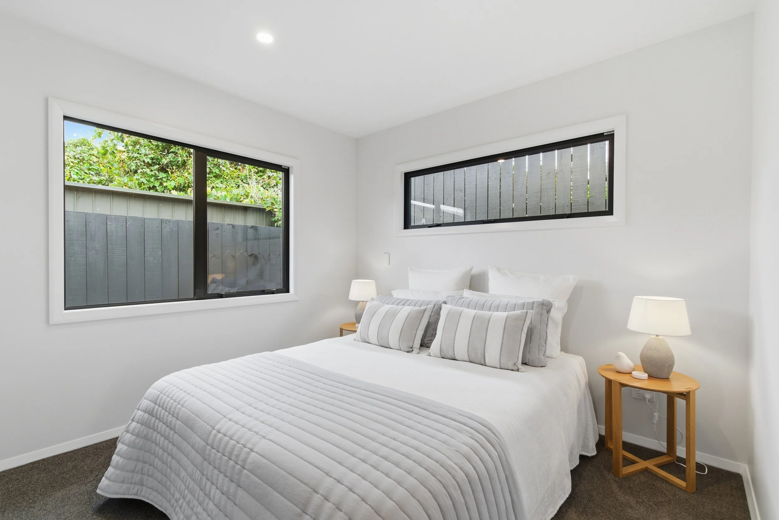 A bedroom with white walls and ceiling, two windows with black frames, a bed with white bedding and striped pillows, a wooden nightstand with a beige table lamp and decorative item, and a gray carpeted floor. New build in Tauranga by BCN Studio