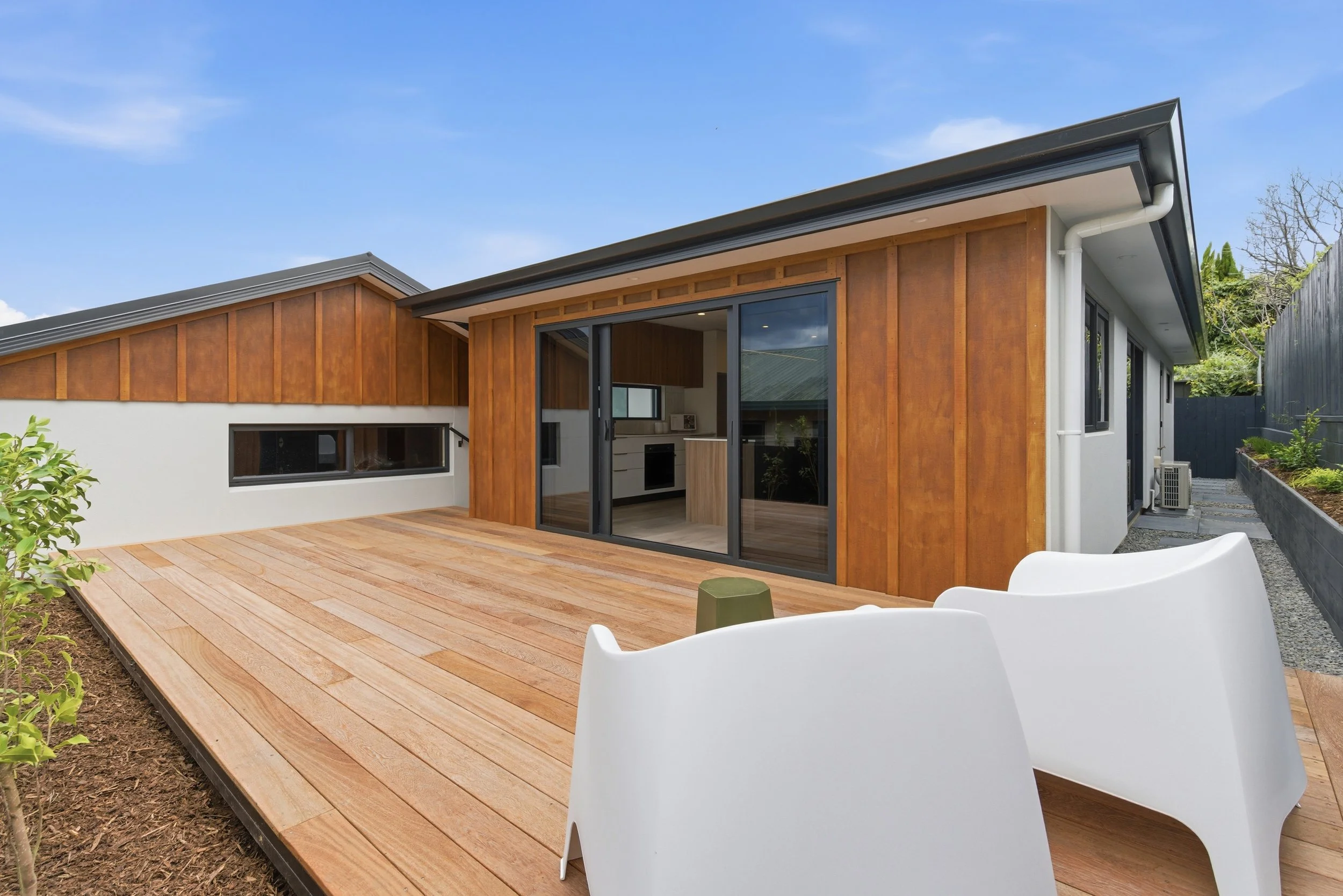 Modern house with a wooden deck and large sliding glass doors, white and wooden exterior, outdoor seating, and a blue sky. New build in Tauranga by BCN Studio