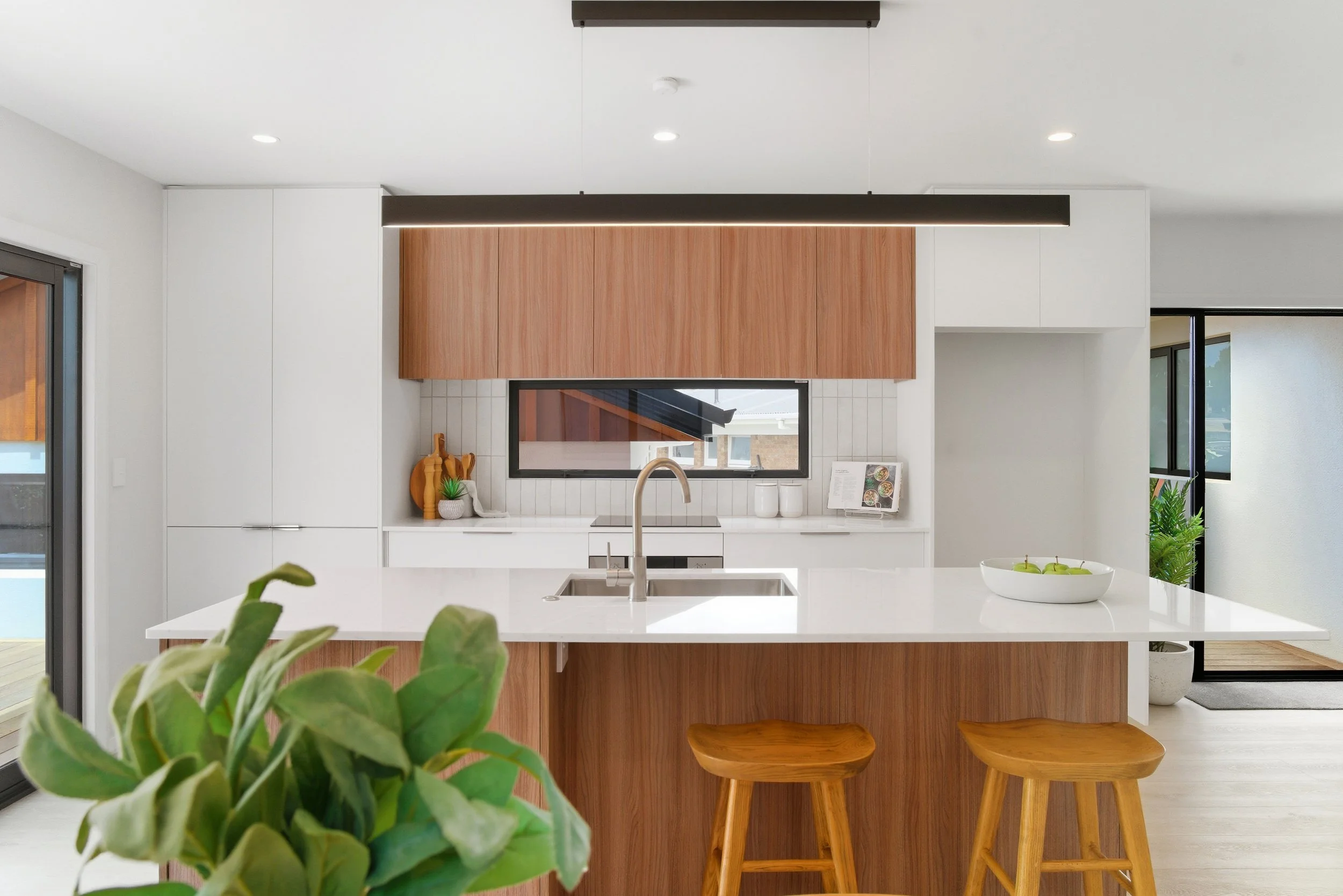 Modern kitchen with white cabinets, wooden accents, a white island with stool, and green plants near a large window. New Build in Tauranga by BCN Studio