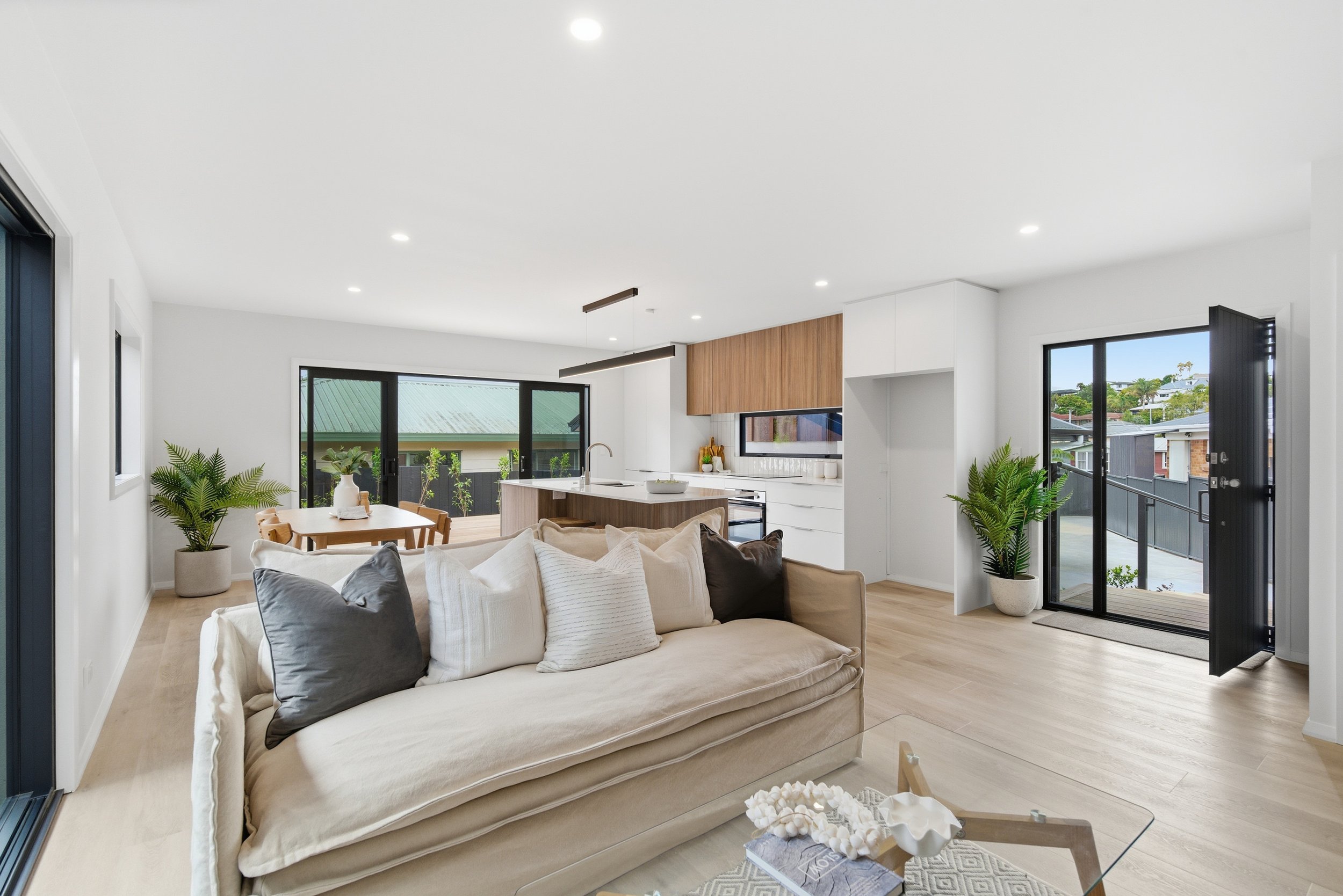 Bright living room with white walls, beige sofa with gray and white pillows, large windows and doors leading to a balcony, modern kitchen with wooden cabinetry and a white island. New build in Tauranga by BCN Studio