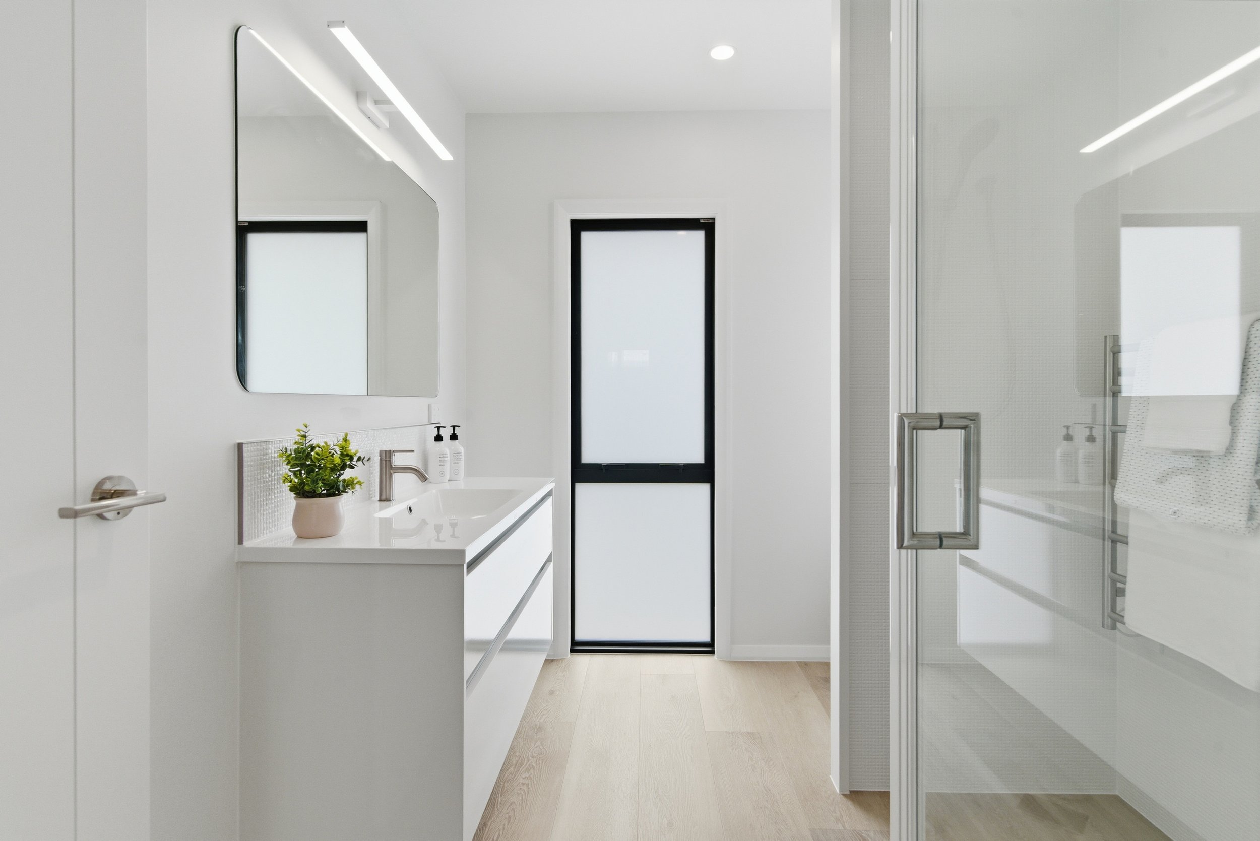 Modern minimalist bathroom with white vanity, large mirror, black-framed window, and glass shower enclosure. New build in Tauranga by BCN Studio
