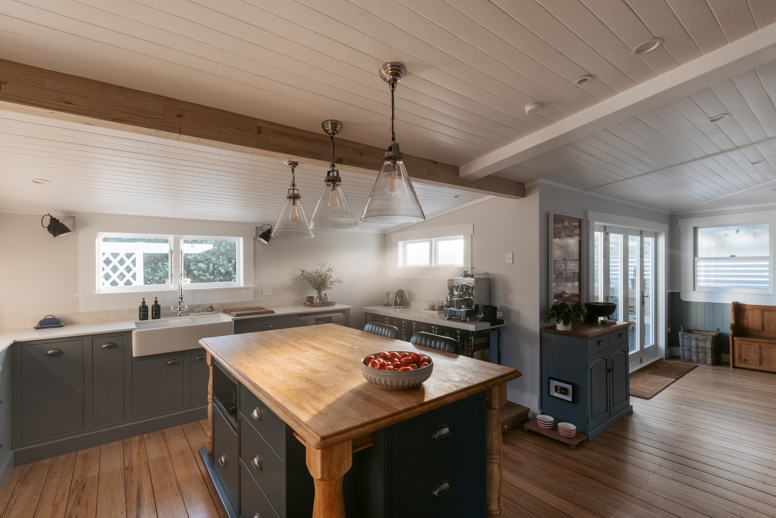 A modern kitchen with gray cabinets, a wooden island with a bowl of tomatoes, and three pendant lights hanging from a white shiplap ceiling. There are windows allowing natural light and a sliding glass door in the background.