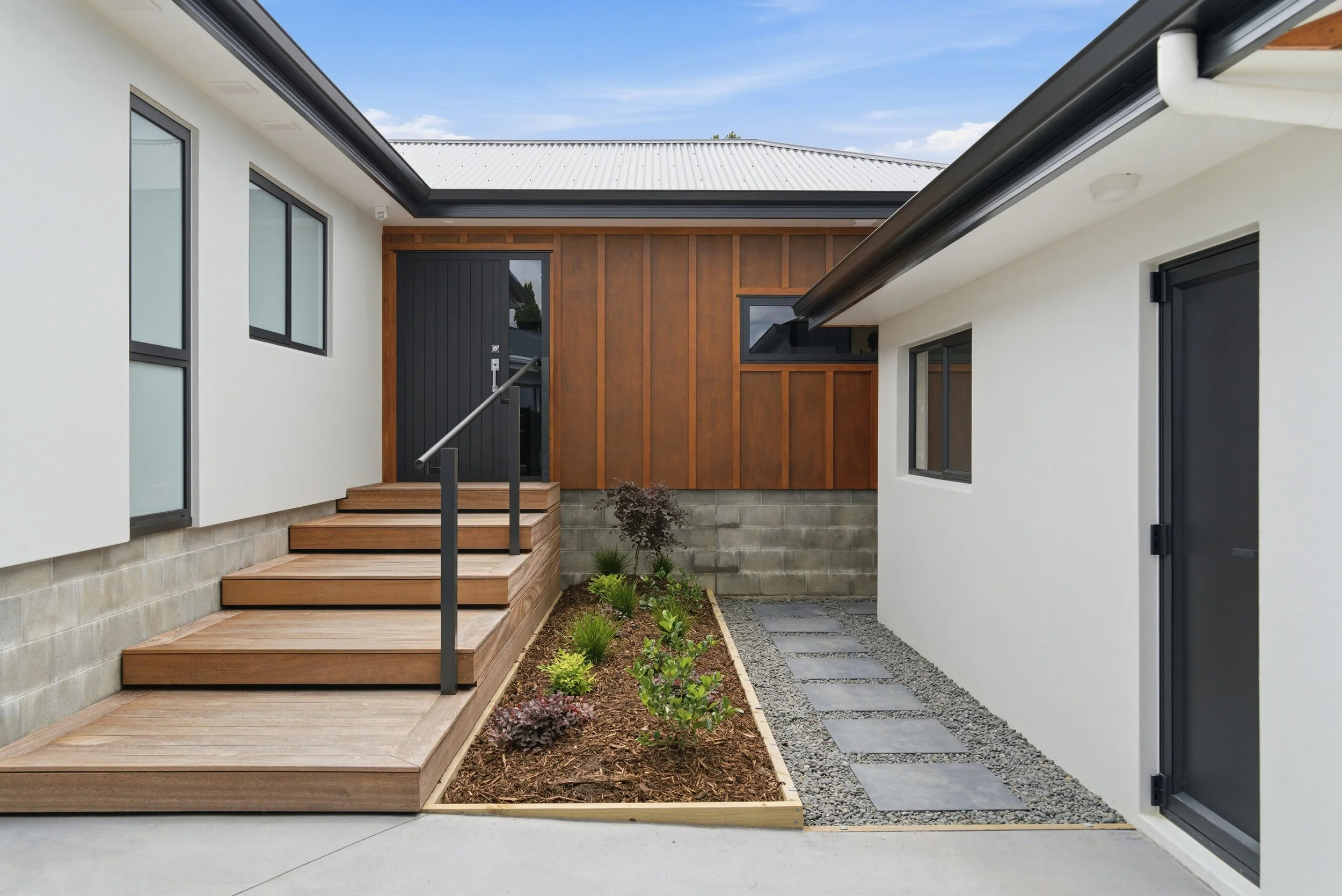 Modern house exterior with white walls, black-framed windows, wooden accents, and a small landscaped garden with stepping stones. New build in Tauranga by BCN Studio