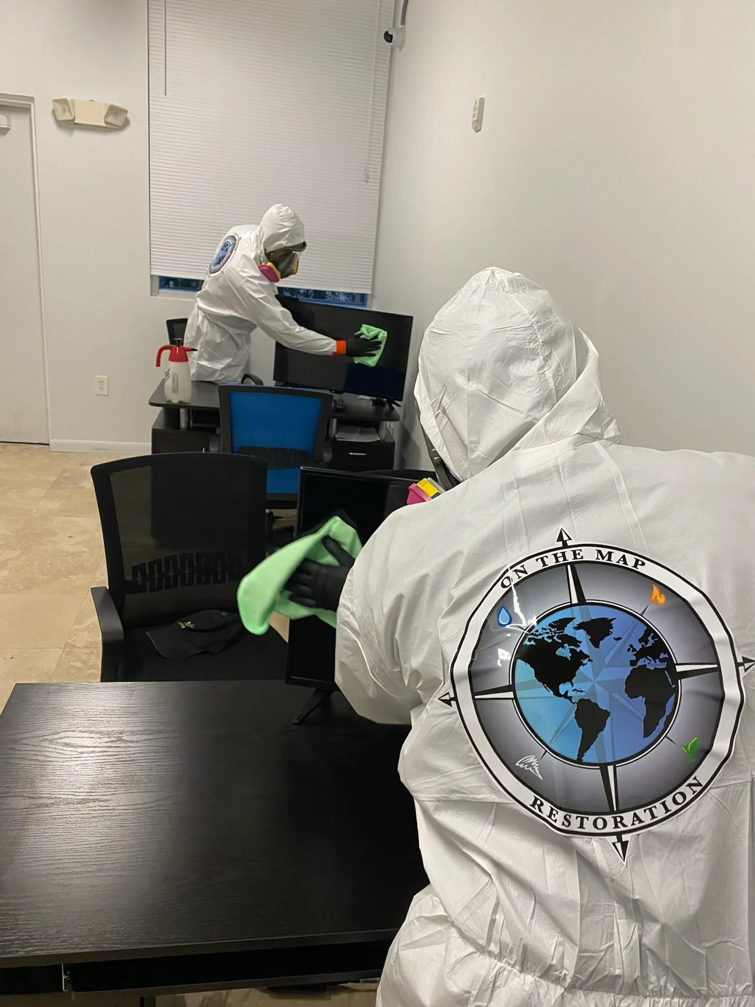 Two OTM technicians in protective suits wiping office surfaces with cloths.