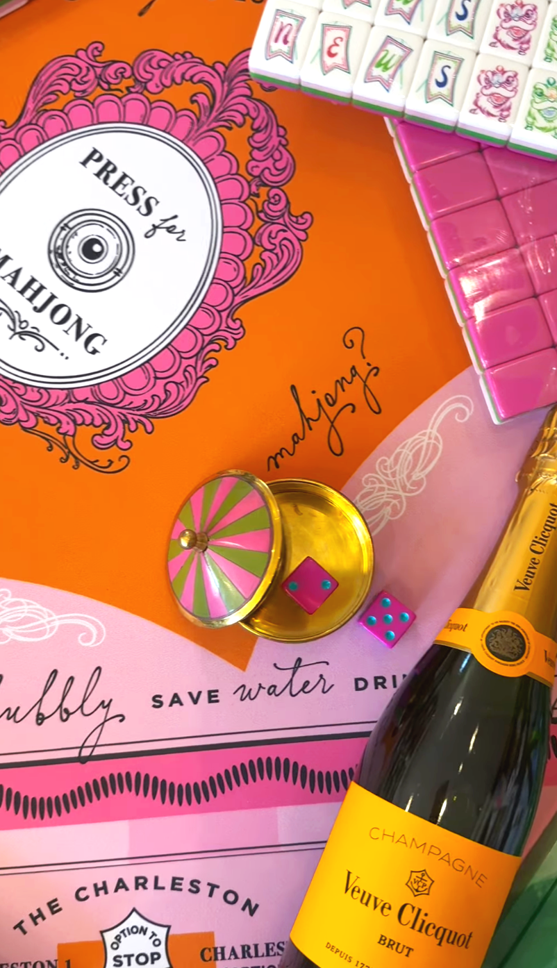 Colorful mahjong board featuring a pink and white sign that says "Press for Mahjong" with decorative pink scrollwork. Displays pink and green dice, pink tiles, a small wheel with pink and green segments, and a bottle of Veuve Clicquot champagne. The 