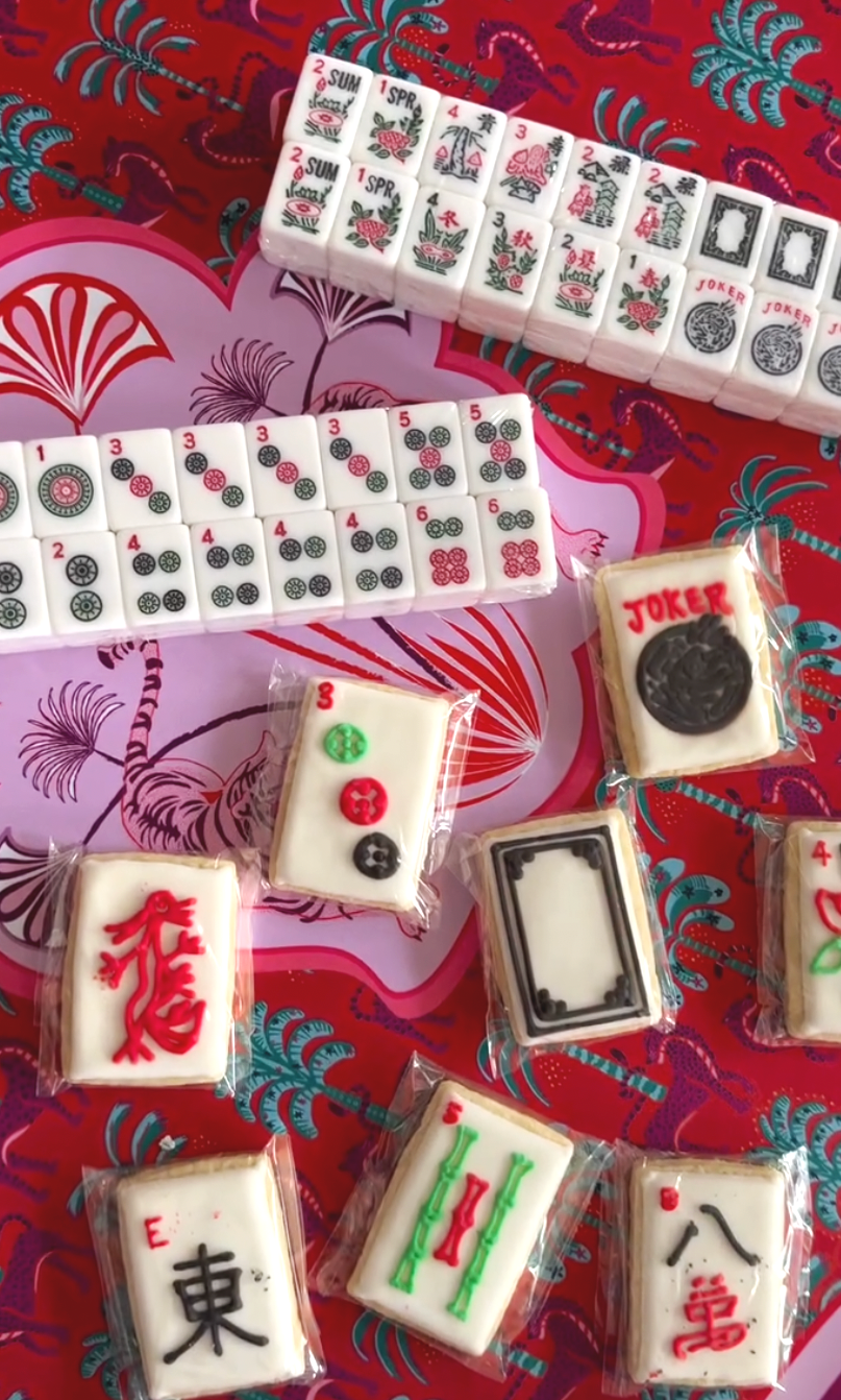 Decorative Mahjong tiles and cookies with Mahjong-themed designs on a red and pink patterned surface.
