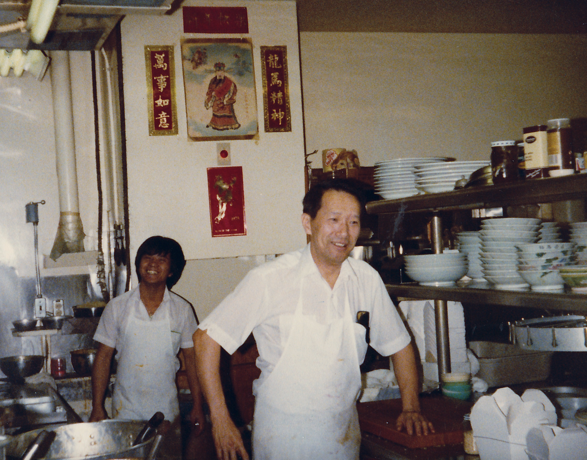 Two chefs in white uniforms standing in a kitchen with stacks of plates and various kitchen supplies behind them. The image features a decorated wall with Asian art and banners.