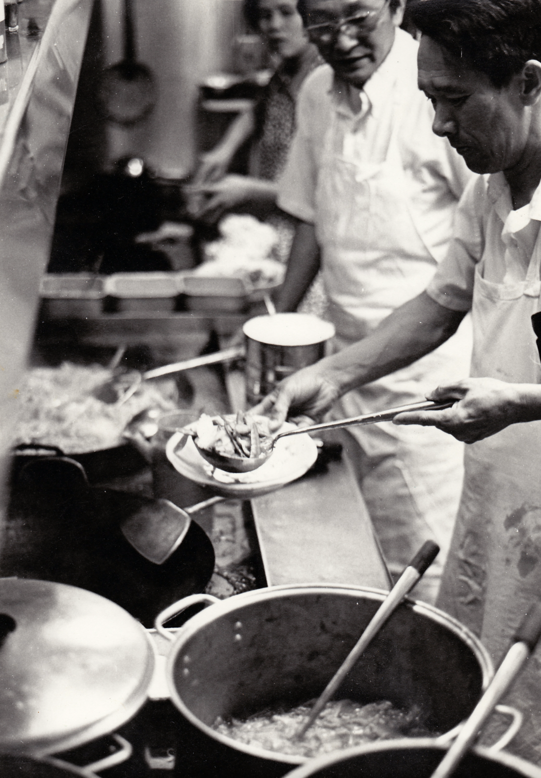 People preparing and serving food in a kitchen, with pots and pans on the stove.