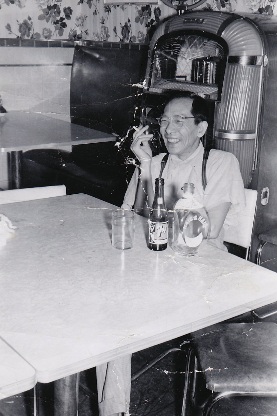An older man with glasses smiling and talking on a rotary phone while sitting at a table with bottles and glasses, in a retro-style room with floral wallpaper.