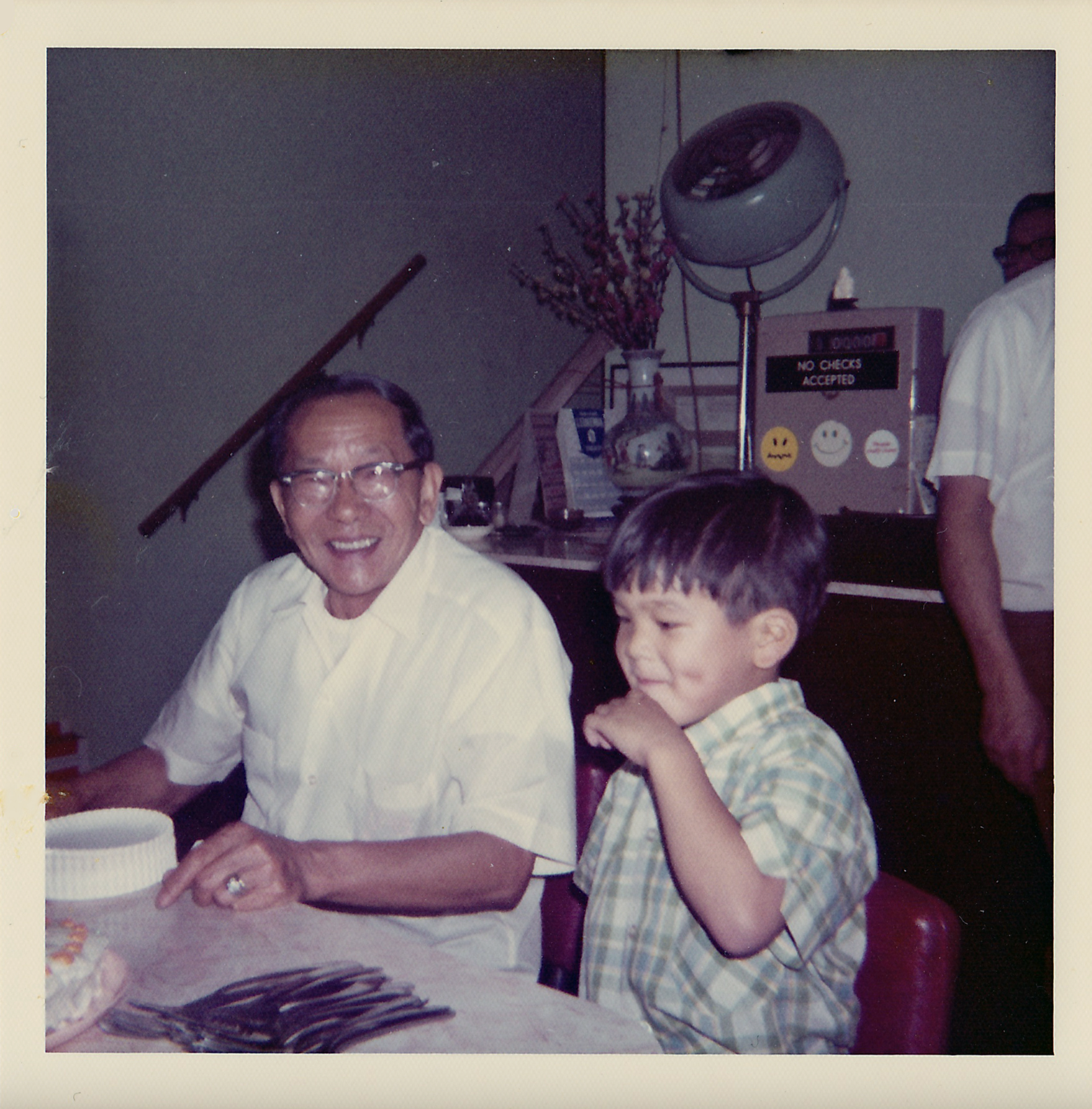 Two men and a boy sitting at a table with a birthday cake, partying in a restaurant or banquet hall with a fan, a telephone, and a sign that says 'No checks accepted' in the background.