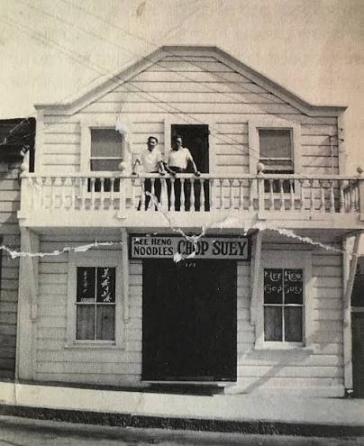 A black-and-white photo of a two-story wooden house with two men standing on the front balcony. The house has a gabled roof, two windows on the lower level, and two windows on the upper level. A sign reading 'Chop Suey' is visible on the balcony railing.