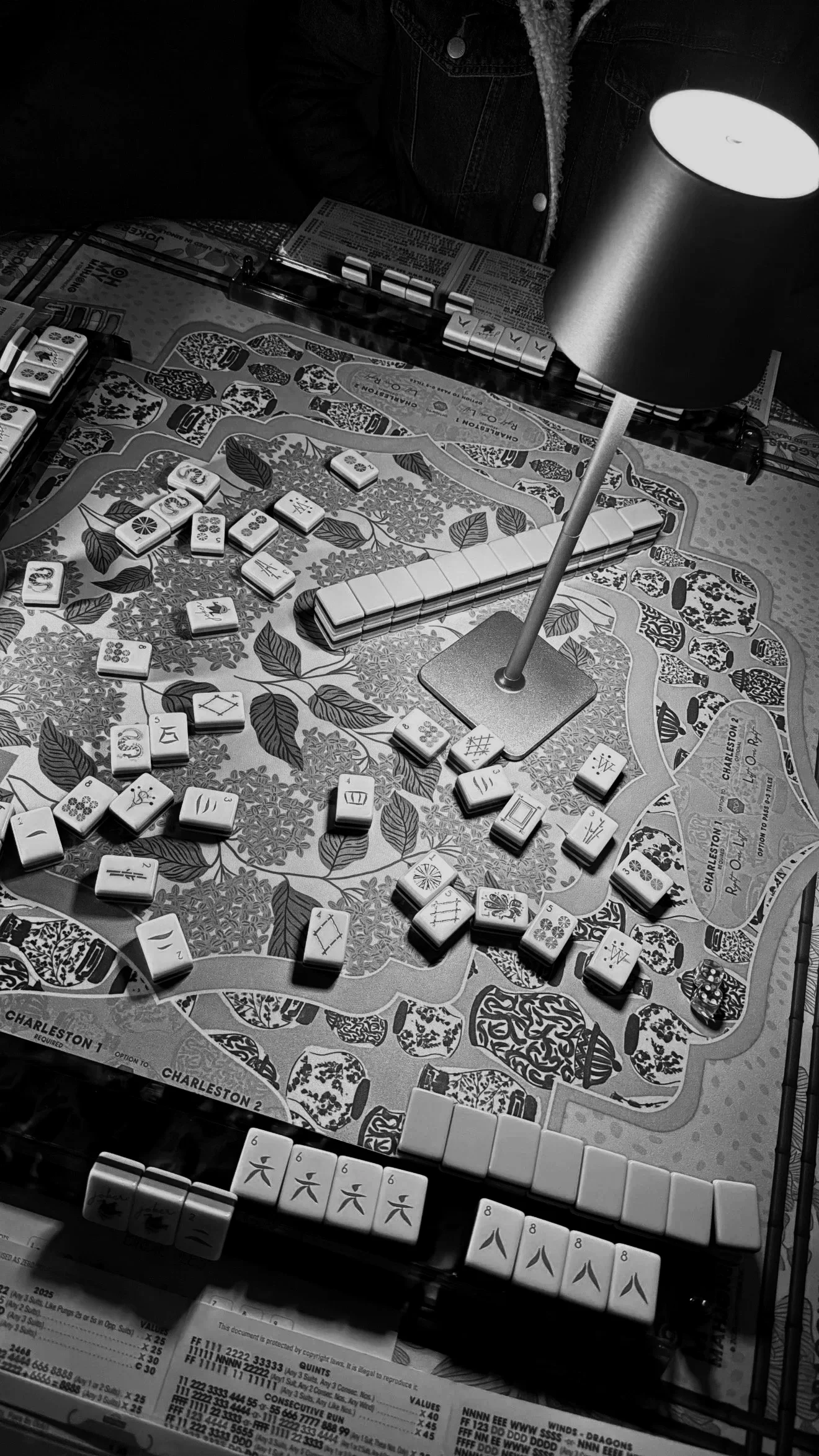 Black and white photo of a board game in progress, featuring various domino tiles, a lamp, and game instructions on a paper at the bottom.