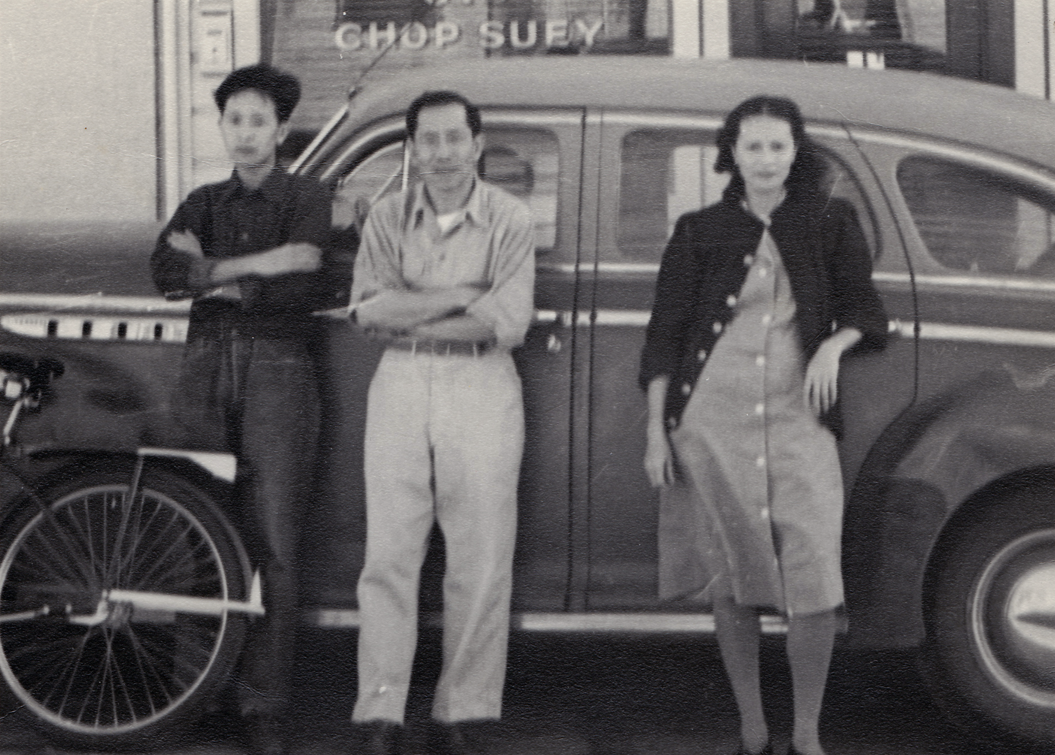 Black-and-white photo of three people standing in front of a vehicle with a sign that says "CHOP SUEY". Two women and one man, dressed casually, posing for the photo.