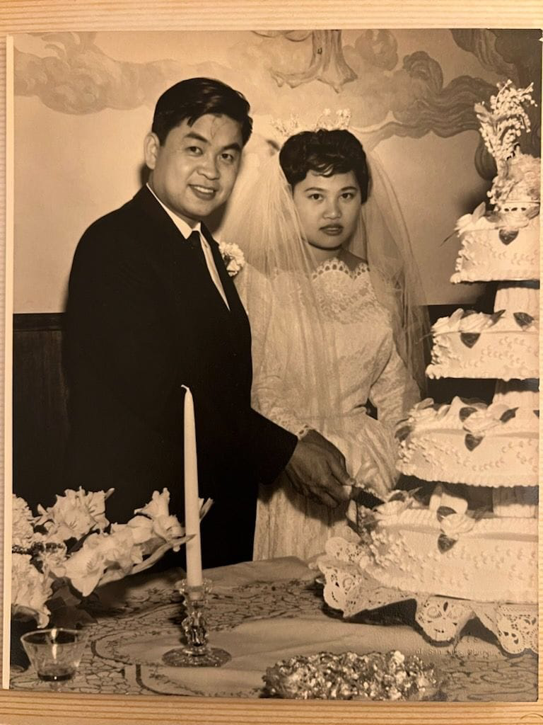 A black and white photo of a bride and groom cutting a multi-tiered wedding cake at their wedding reception. The groom is wearing a dark suit and tie, while the bride is in a lace wedding dress with a veil.