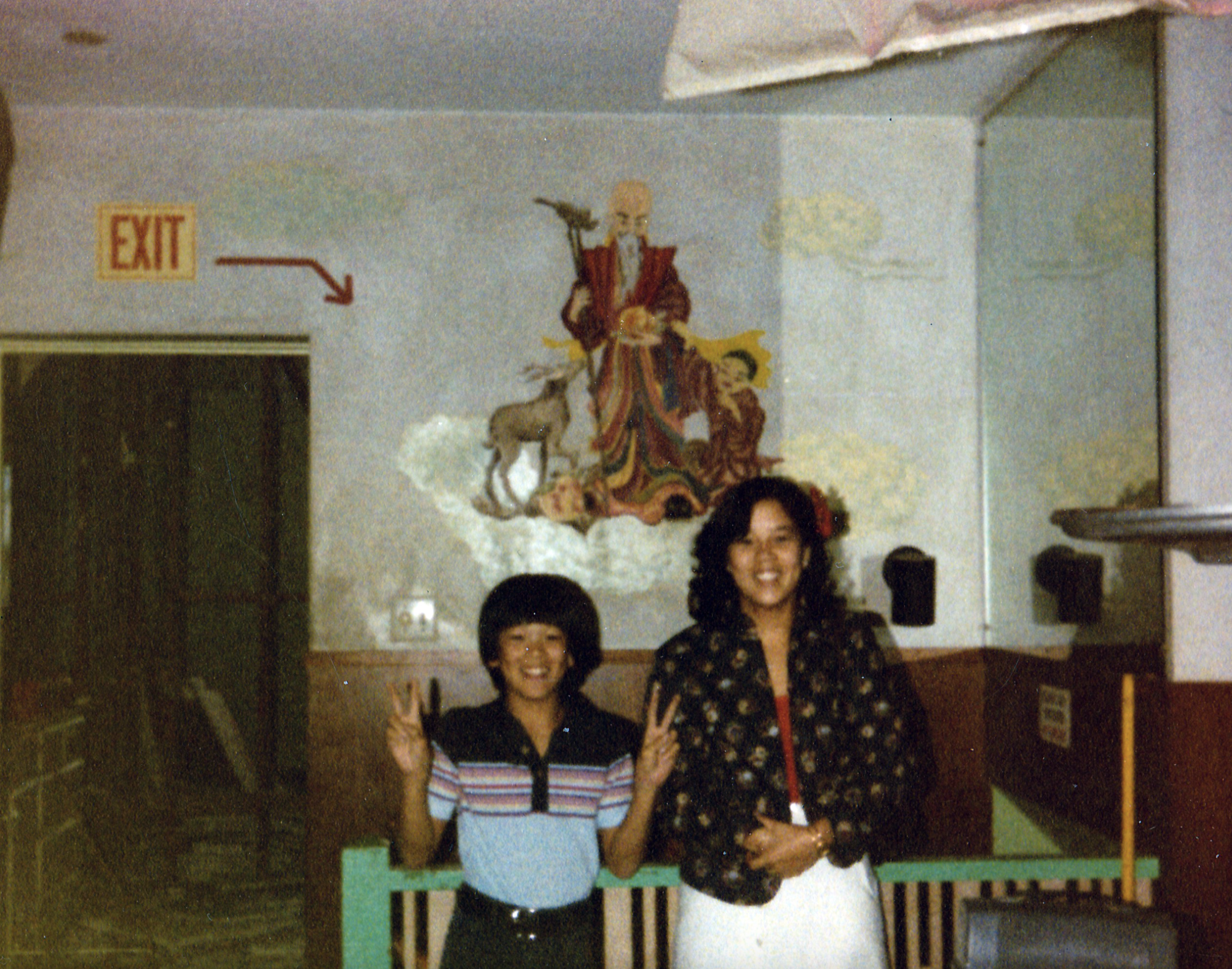 Two people smiling and posing for a photo in what appears to be a decorated indoor space, with one person making a peace sign and the other standing calmly.