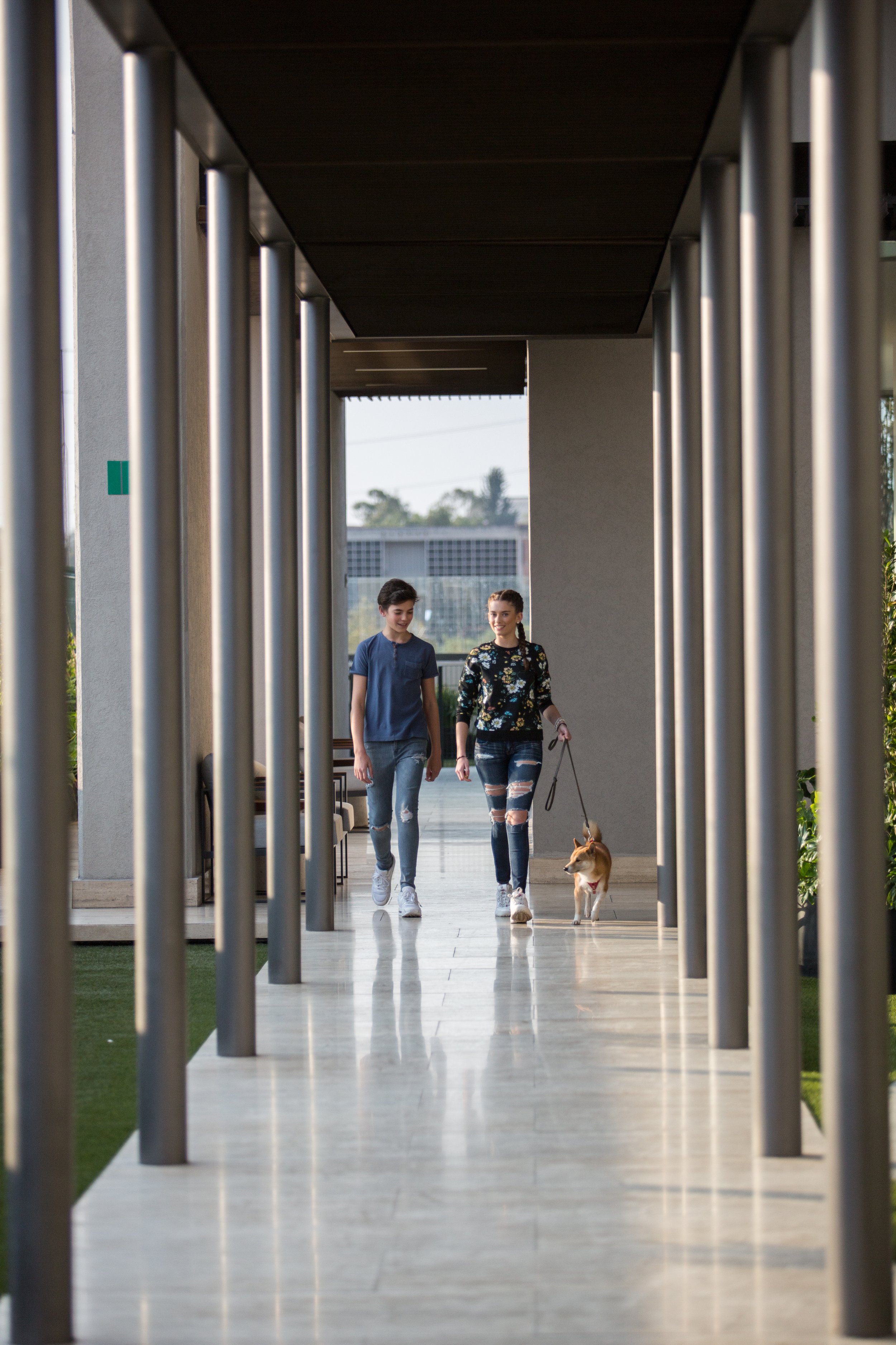 Two women walking with a dog in an outdoor covered walkway with metal poles and a modern building in the background.