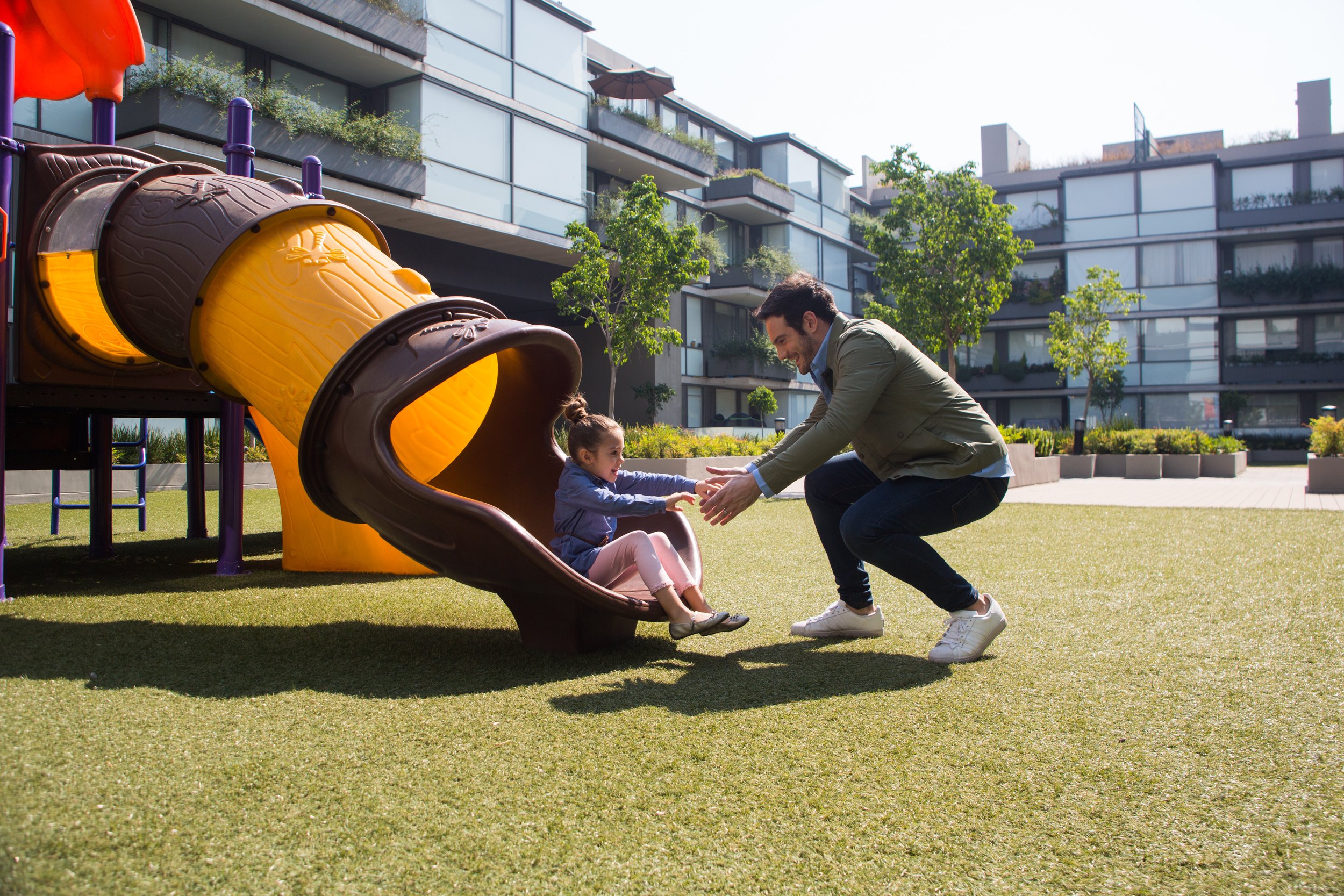 A man and a young girl playing on a playground grass field, with the girl sitting on a slide and the man kneeling and holding her hands, smiling. There are modern apartment buildings and trees in the background.