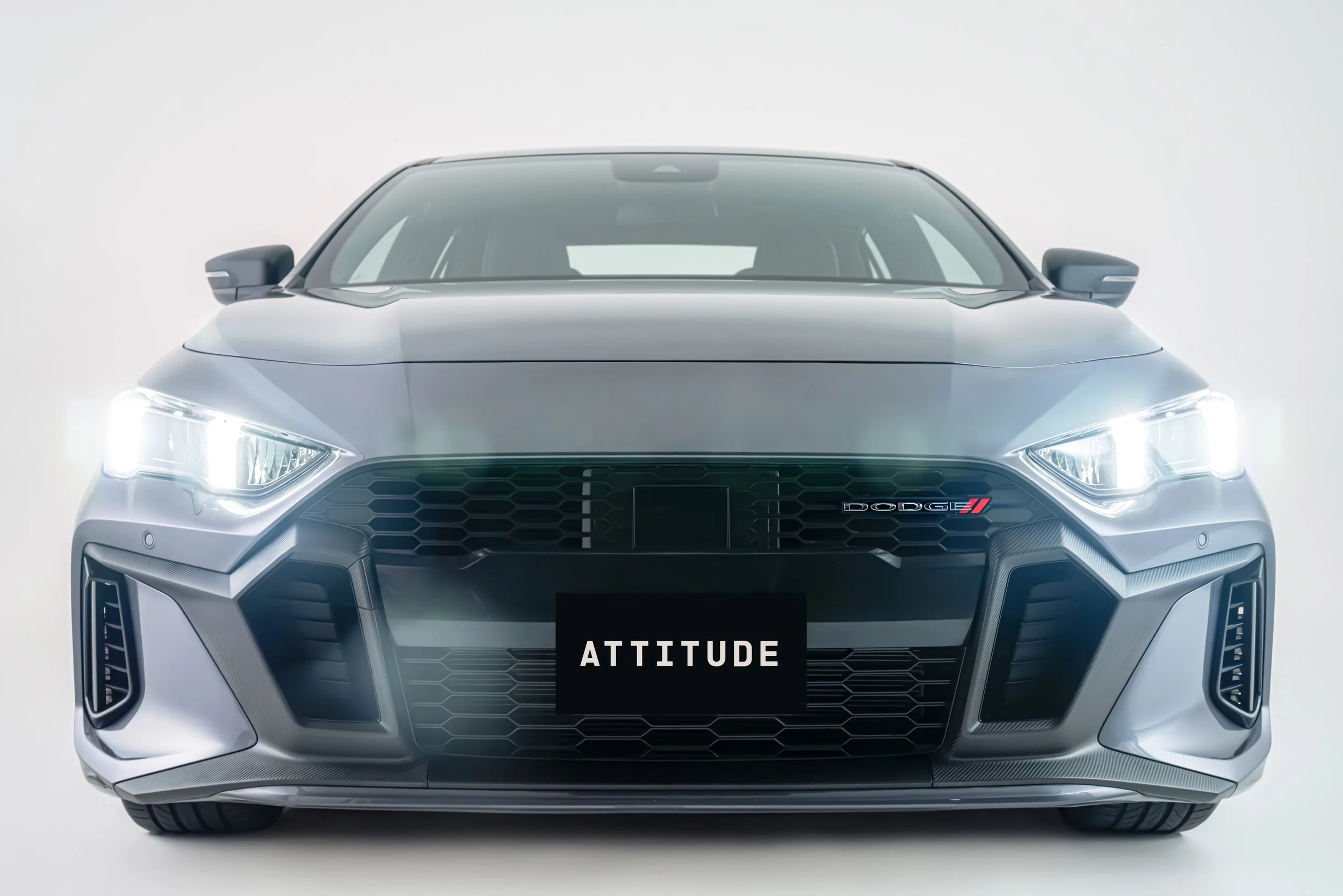 Front view of a silver modern sporty car with LED headlights, a black grille with the Dodge logo and the word ATTITUDE on a black background.