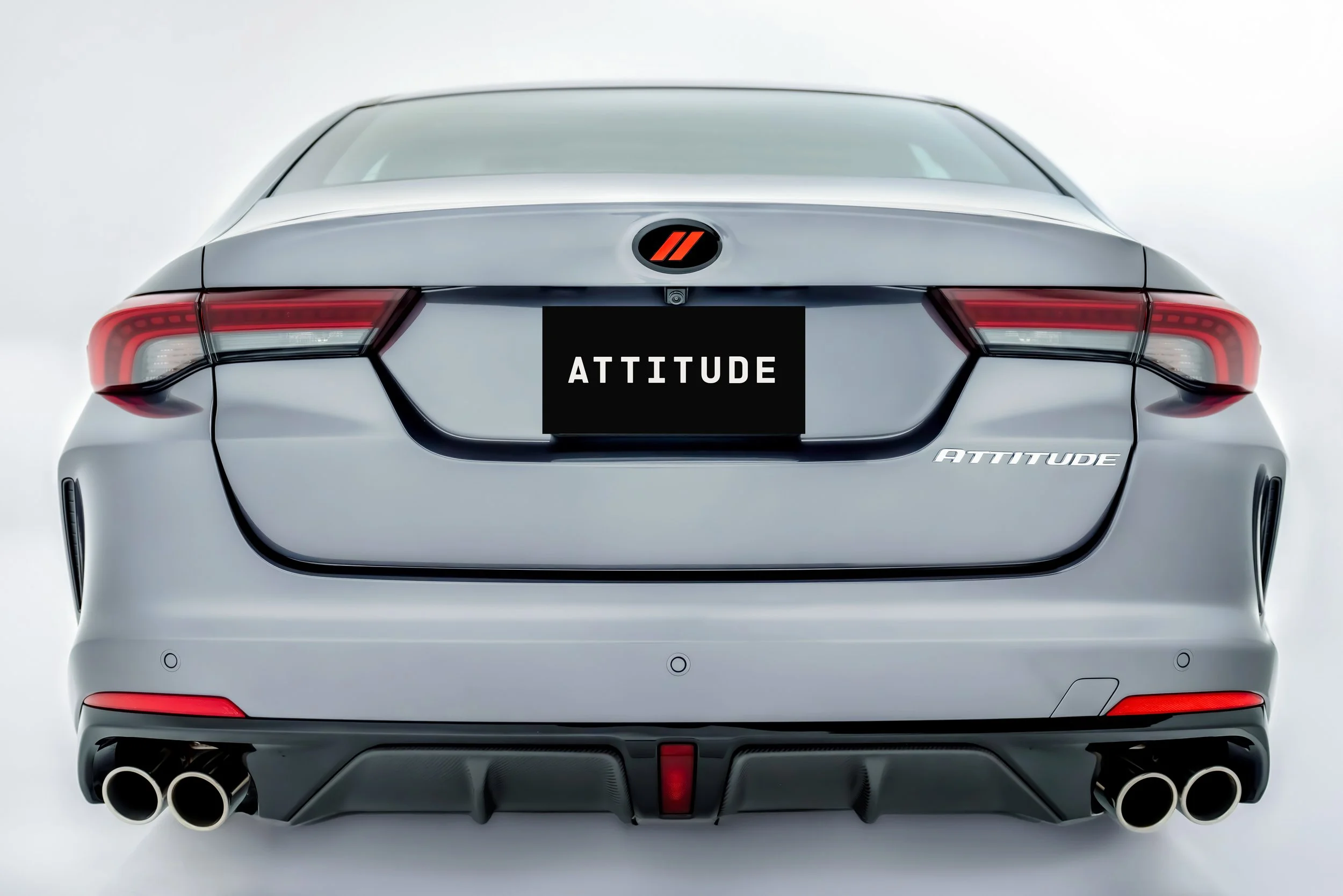 Rear view of a silver car with the word 'ATTITUDE' on the license plate and a logo on the trunk. The car has modern taillights and dual exhaust pipes.