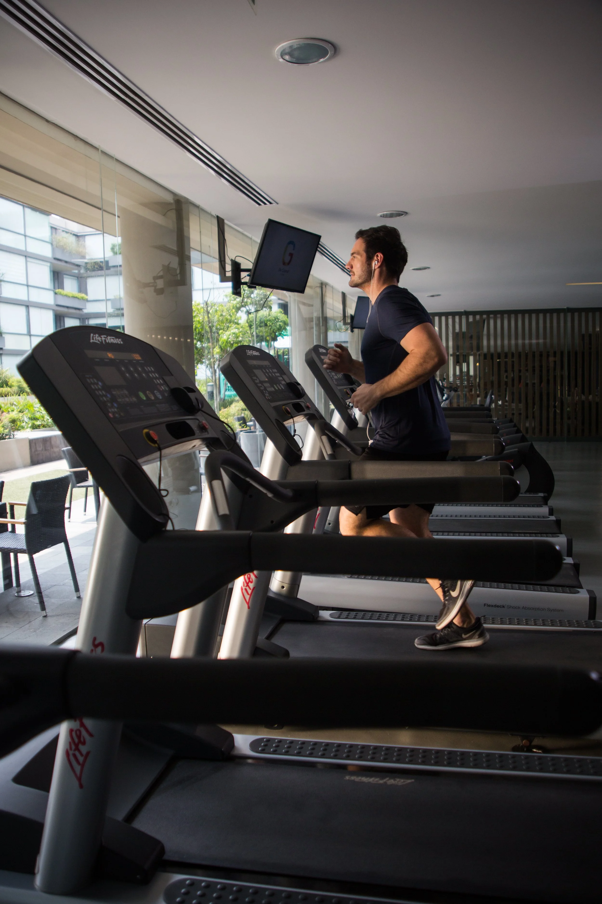 A man running on a treadmill at a modern gym with large windows and a view of buildings and trees outside.