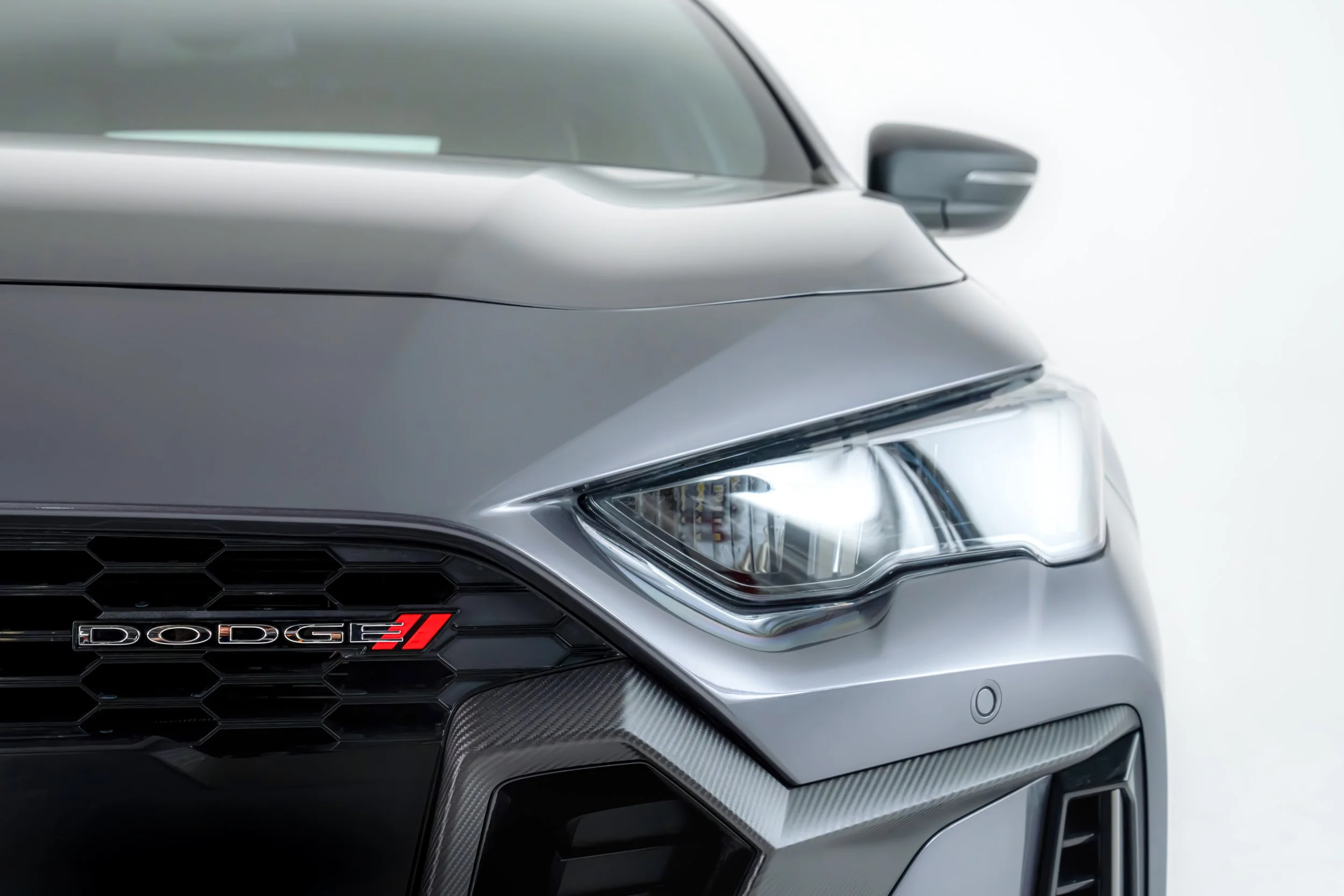 Close-up of the front of a silver Dodge vehicle, showing part of the headlight, grille with hexagonal pattern, and the Dodge emblem with an SRT badge.