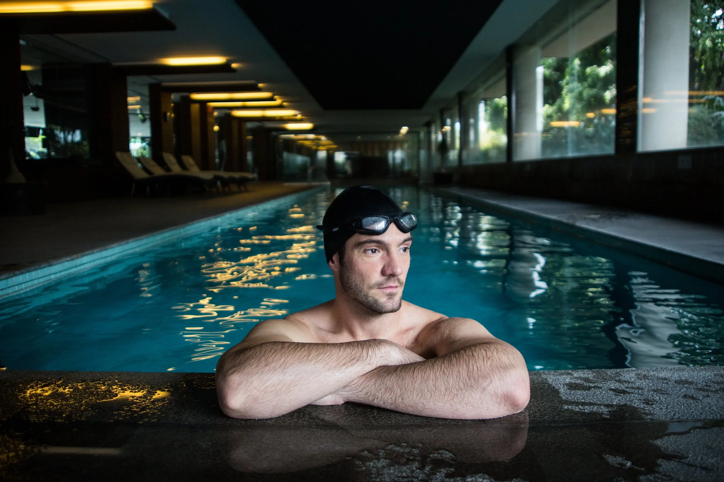 A man in a swimming cap and goggles rests on the edge of an indoor swimming pool, with his arms crossed in front of him, in a modern facility with large windows and seating area.