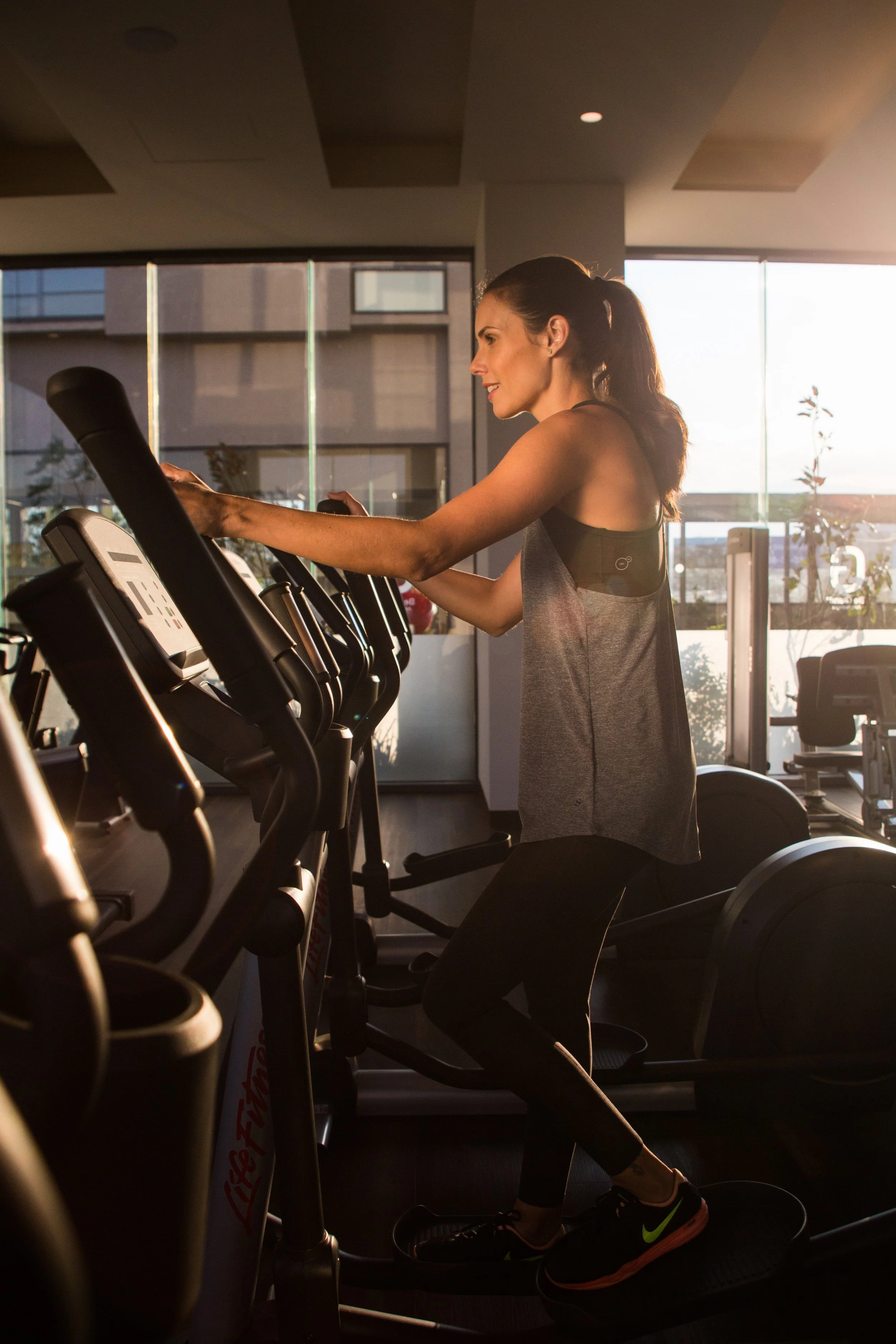 A woman in athletic clothing using a treadmill in a gym during sunset with large windows in the background.