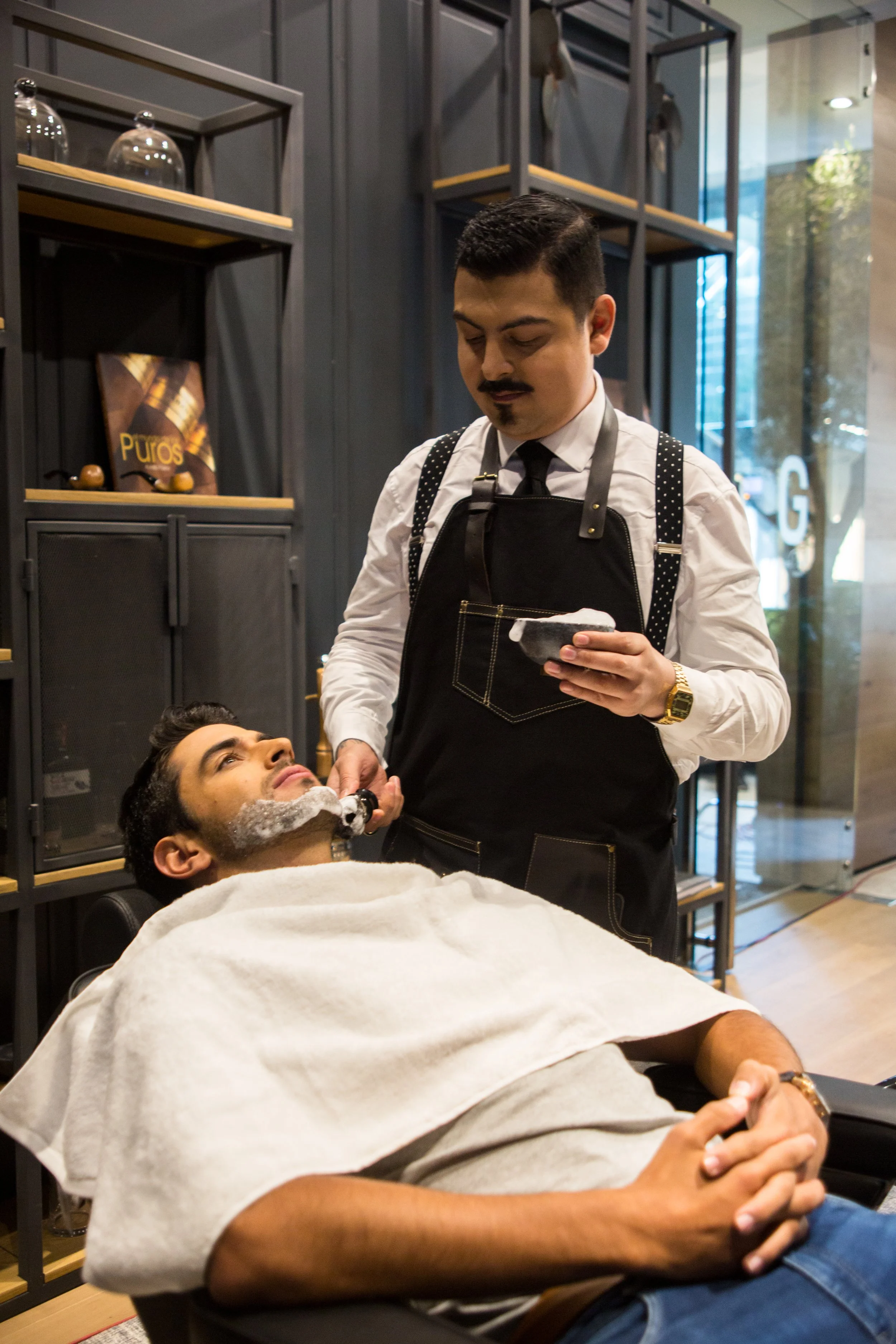 A man with a white towel over his chest is sitting in a barber chair as another man, dressed in a white shirt, black apron, and polka dot suspenders, gives him a shave with a straight razor. The barber is holding a soap bowl, and the man receiving th