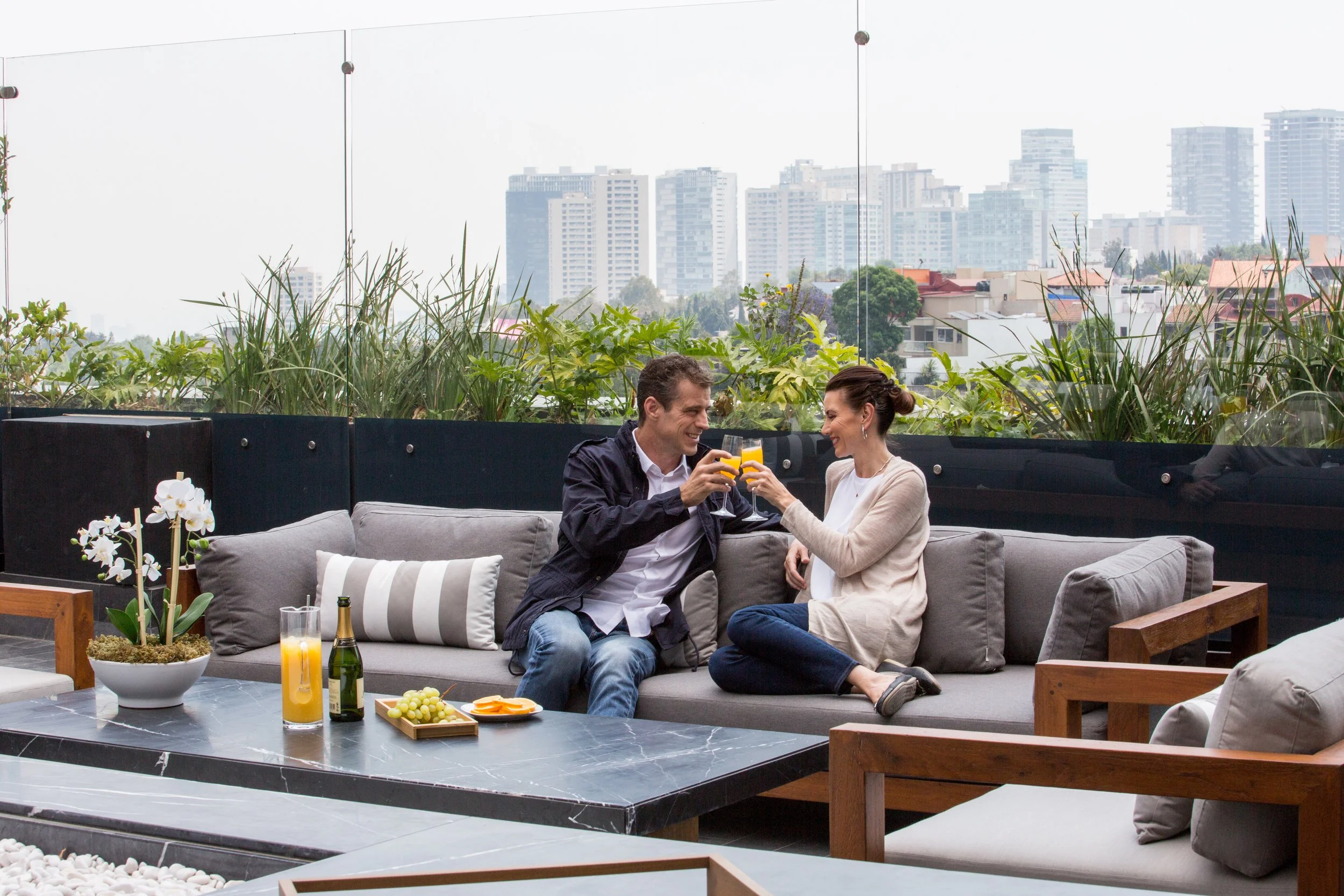 A man and woman sit on an outdoor sofa, smiling and clinking glasses of orange juice. The setting includes a modern rooftop with plants, a table with snacks, and a city skyline background.