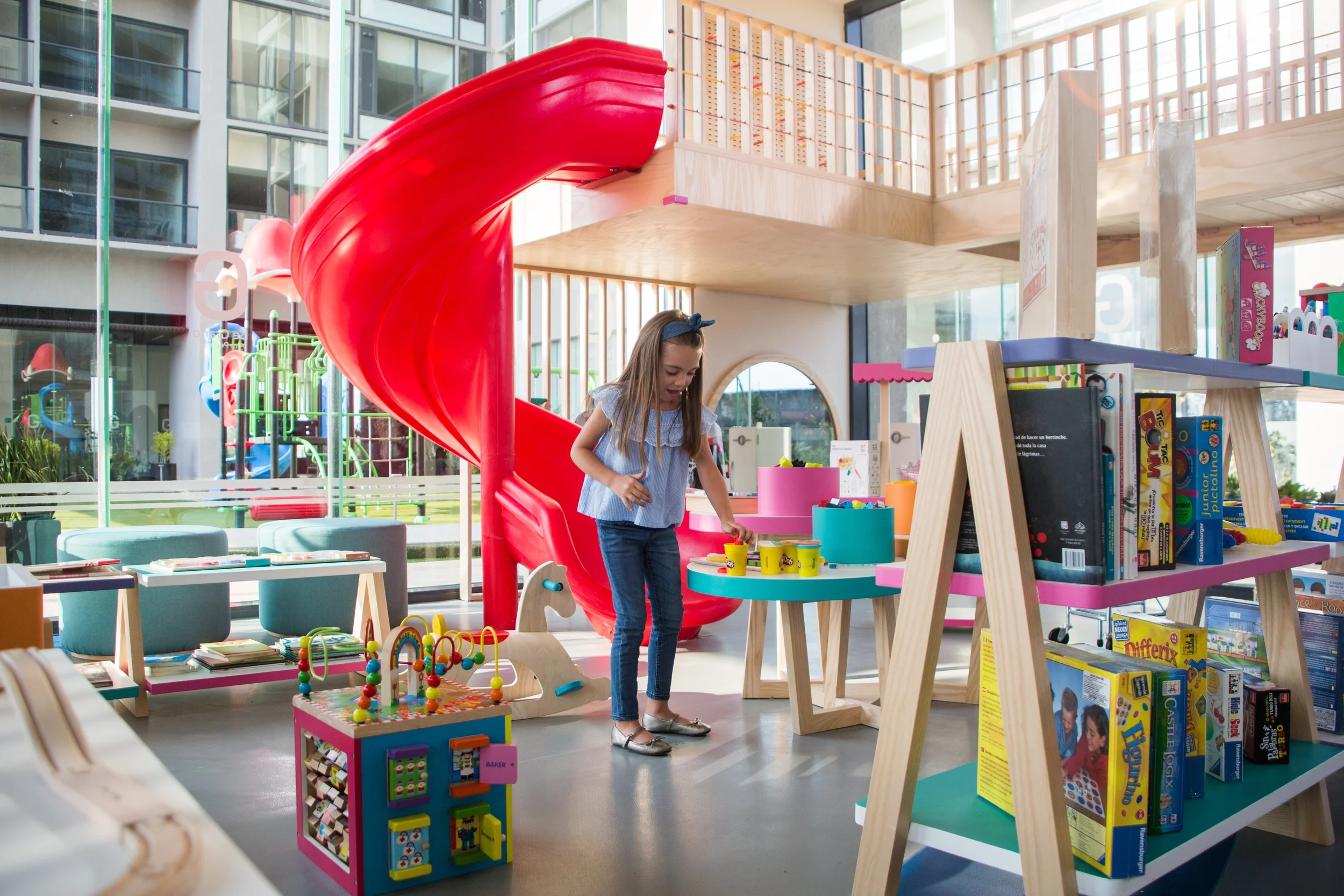 A young girl with a blue bow in her hair is playing with colorful toys in a bright indoor children's play area with a red slide, bookshelves, and large windows.