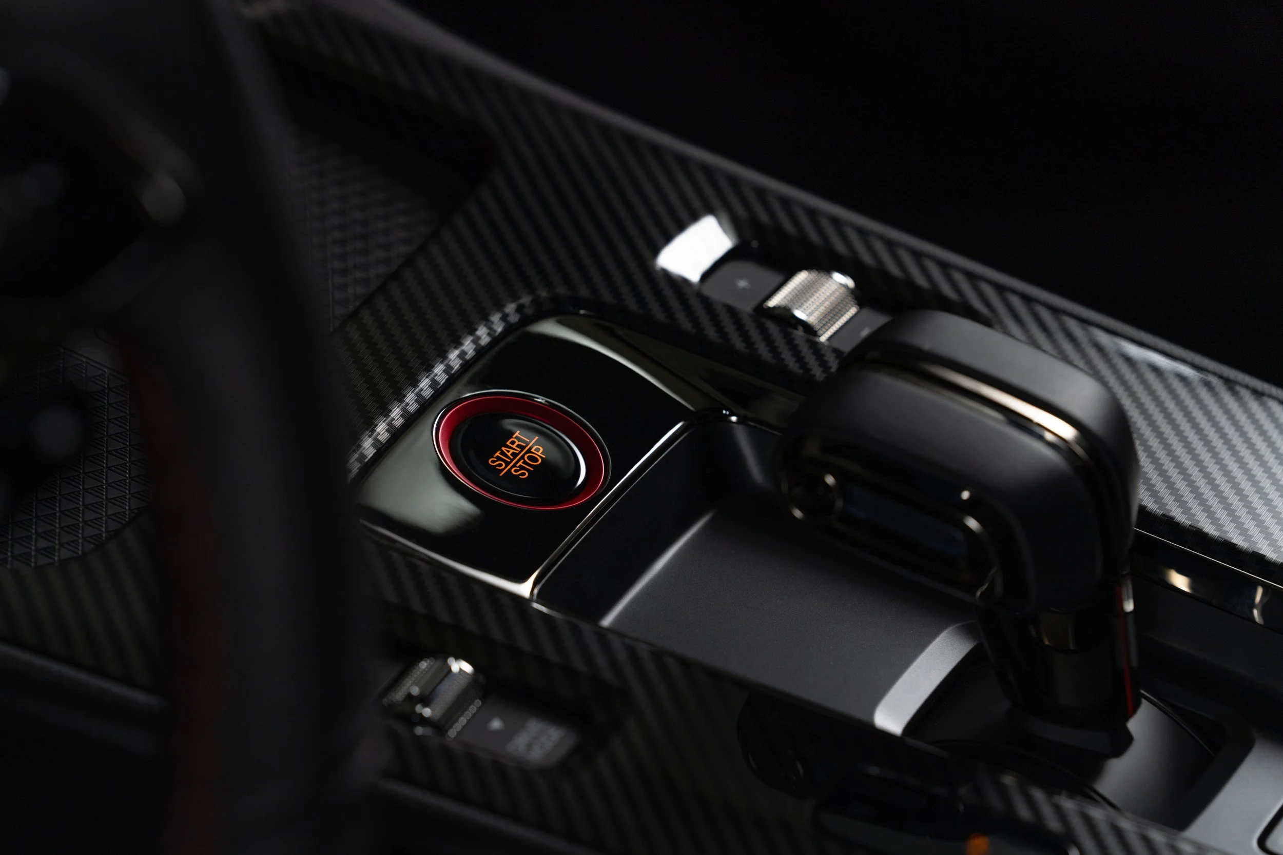 Close-up of a car's interior focusing on a start/stop button with red circle, set in a dashboard with a carbon fiber pattern.