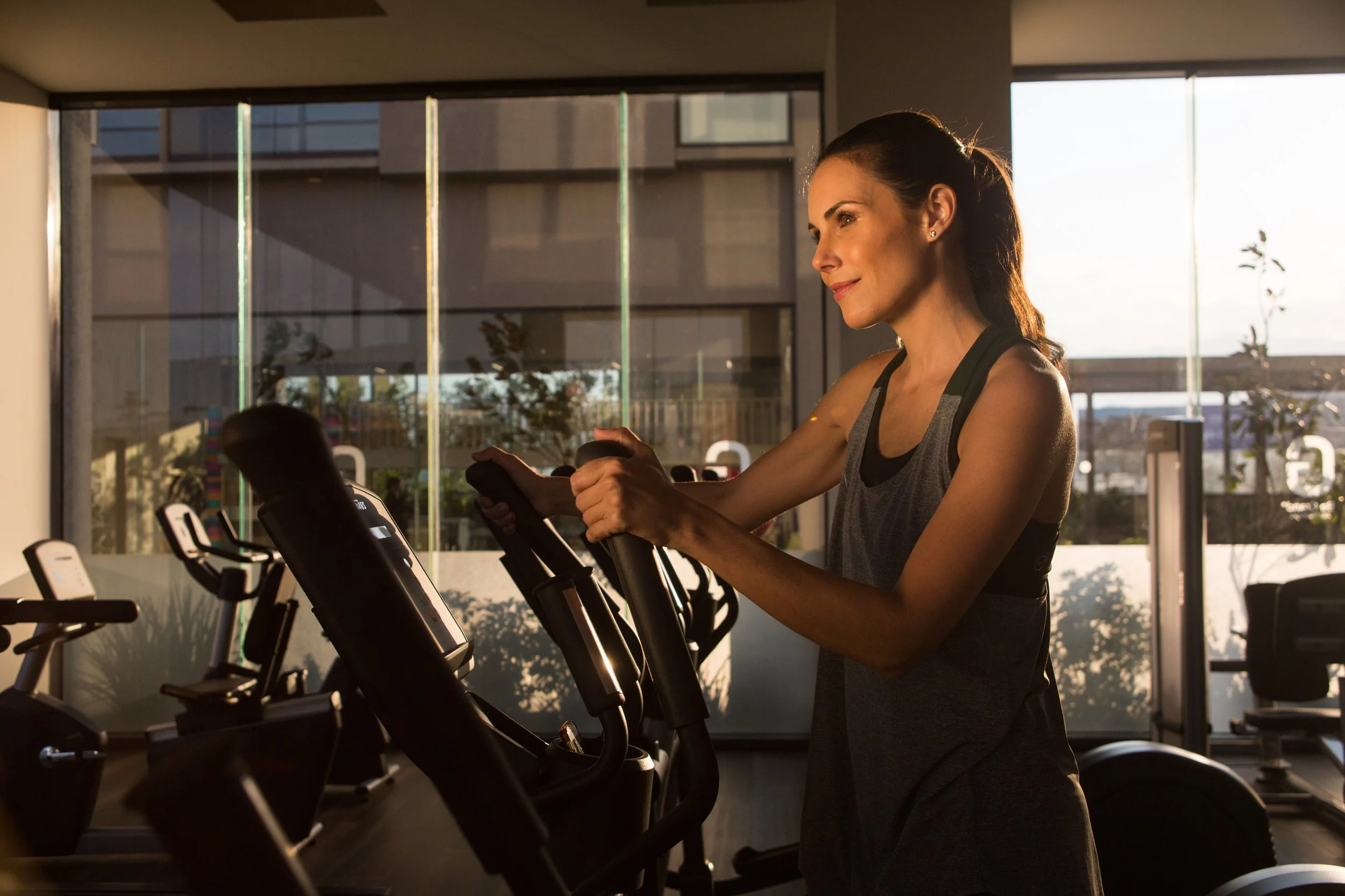 A woman exercising on a stationary bike at the gym, with large windows in the background letting in natural sunlight.