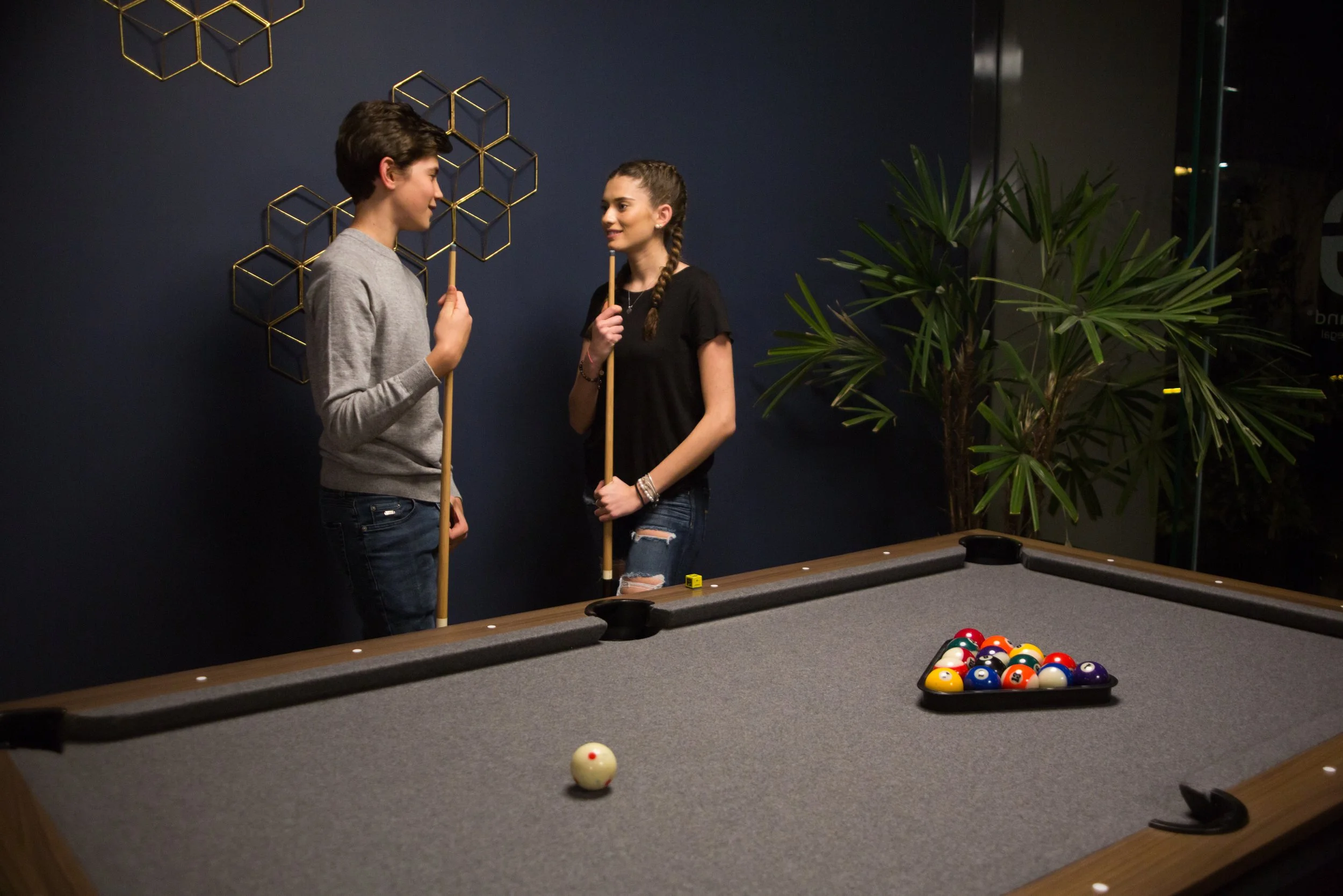 Two young people, a boy and a girl, stand near a pool table holding cue sticks, engaged in conversation, with a rack of pool balls on the table and decorative geometric wall art and a large potted plant in the background.