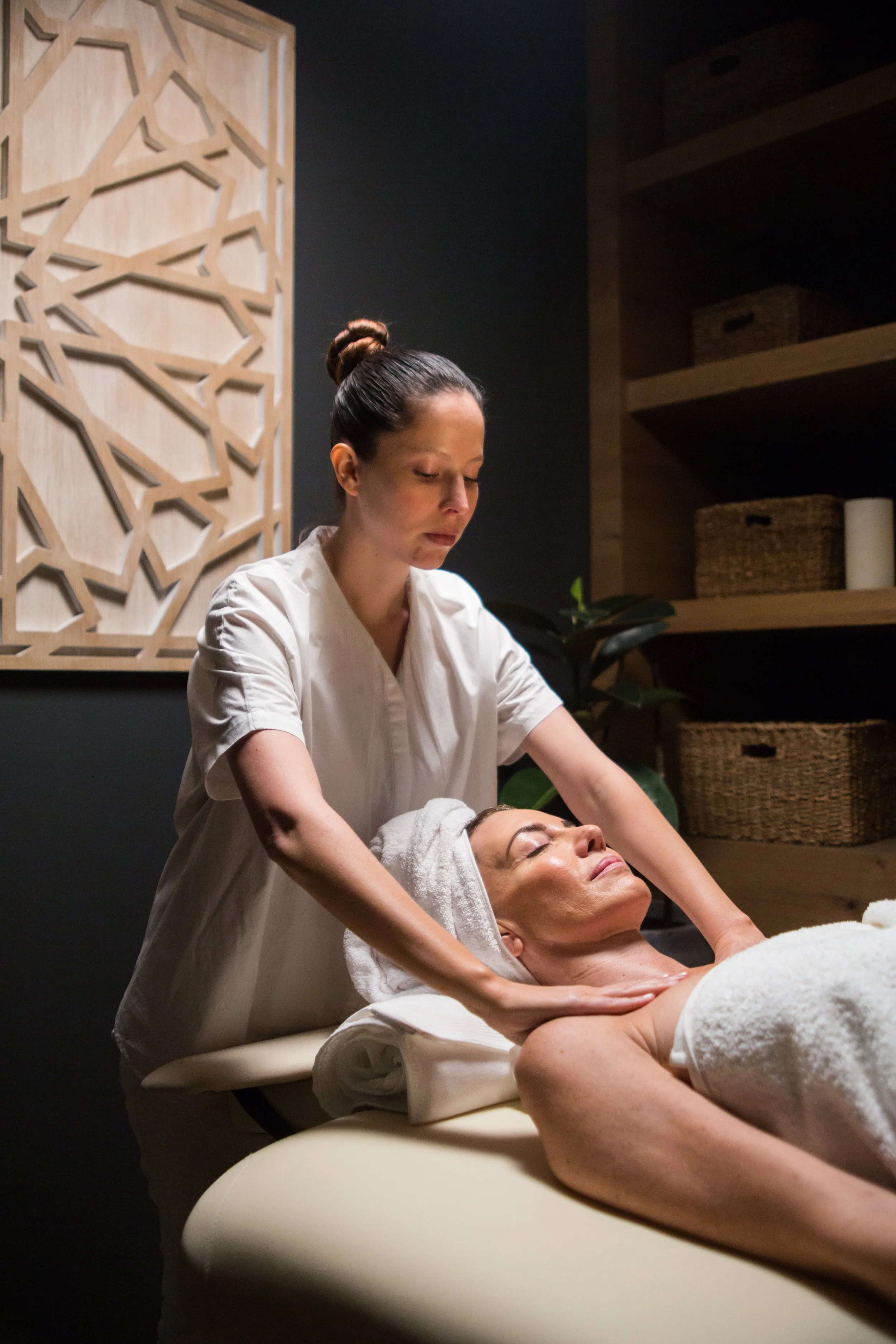 A woman receiving a facial massage from a spa therapist in a wellness massage room with wooden shelves and basket storage.
