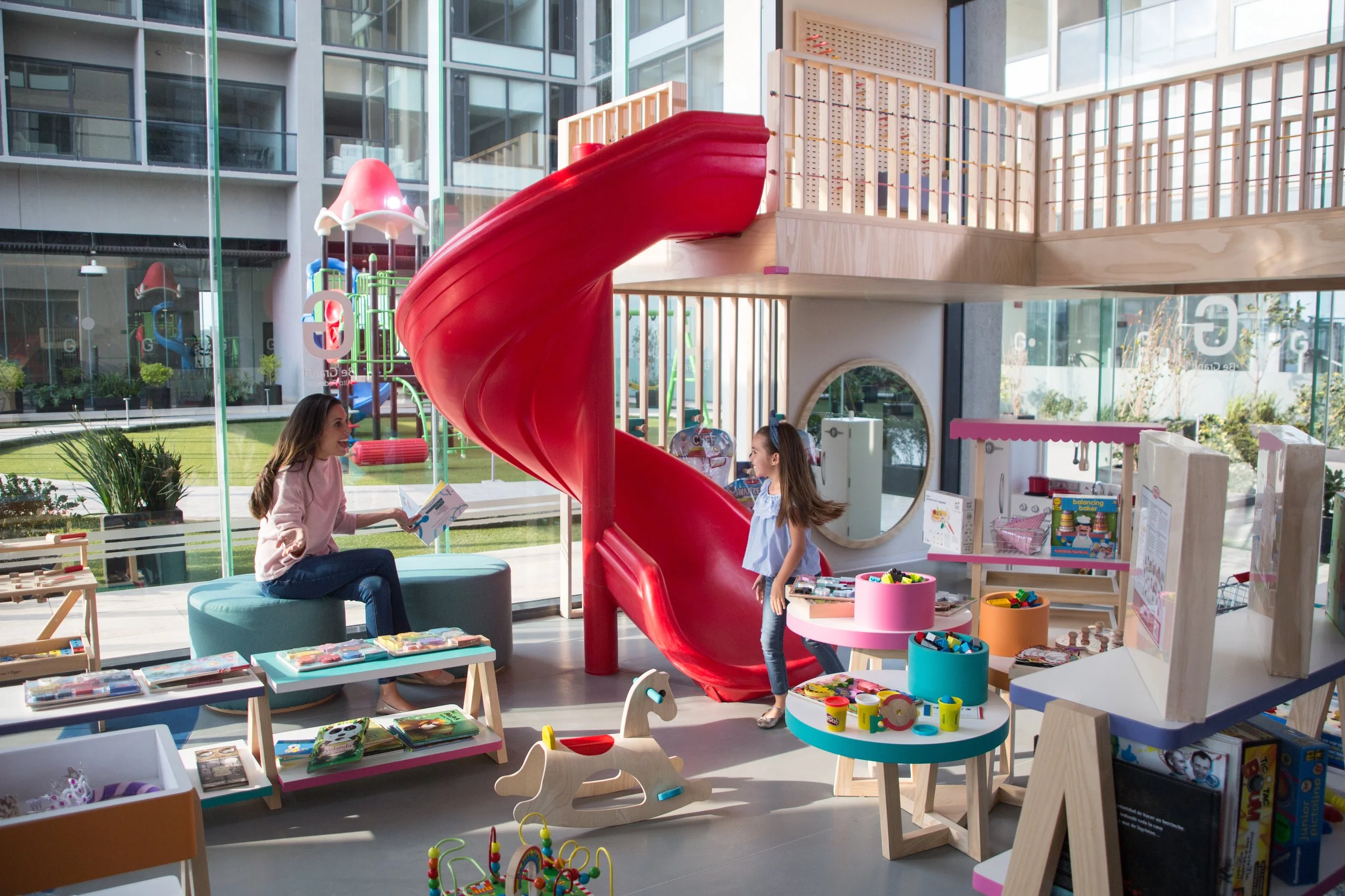 Indoor children's play area with a red spiral slide, toys, books, and a woman sitting on a teal ottoman talking to a girl.