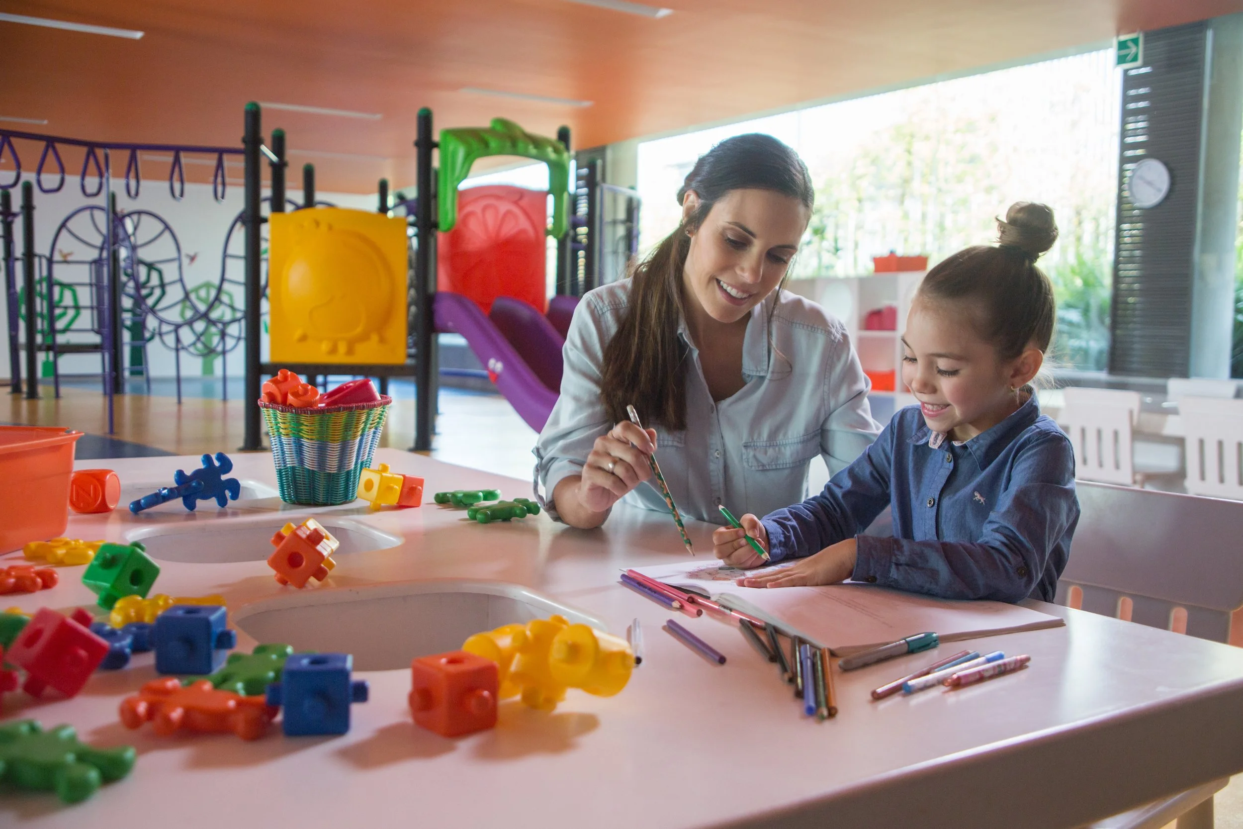A woman and a young girl sitting at a table in a colorful indoor play area, engaging in drawing or coloring activities, with various toys and art supplies scattered on the table.