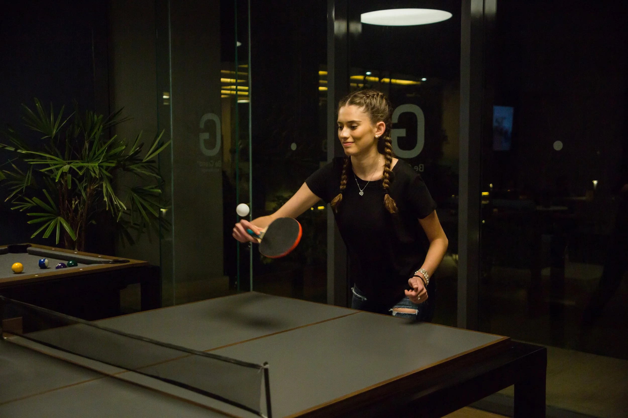 A young woman with braided hair playing table tennis in a dimly lit room with a pool table in the foreground and decorative plants on the side.