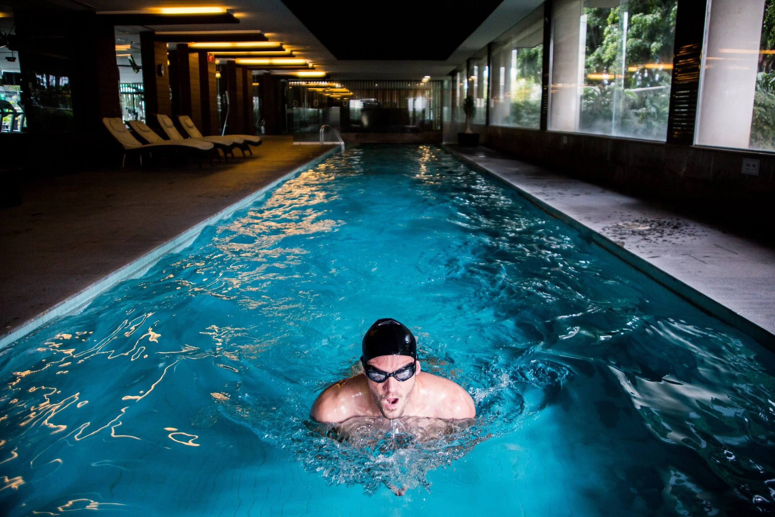 A person swimming in an indoor pool, wearing goggles and a swim cap, with sunlight coming through large windows.