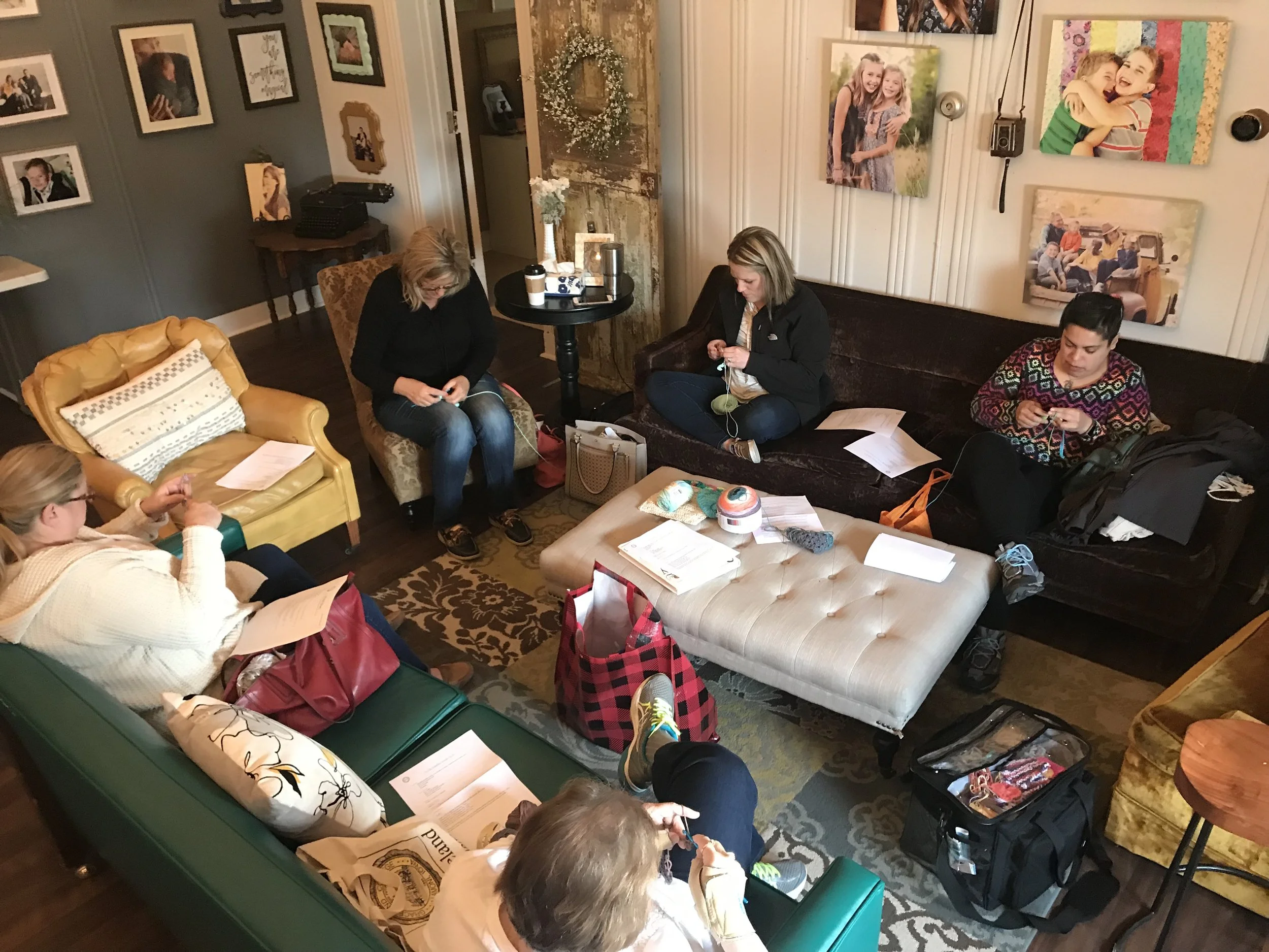 A group of people sitting in a living room, knitting and reading papers, with photos on the walls, a lamp, and personal belongings around.